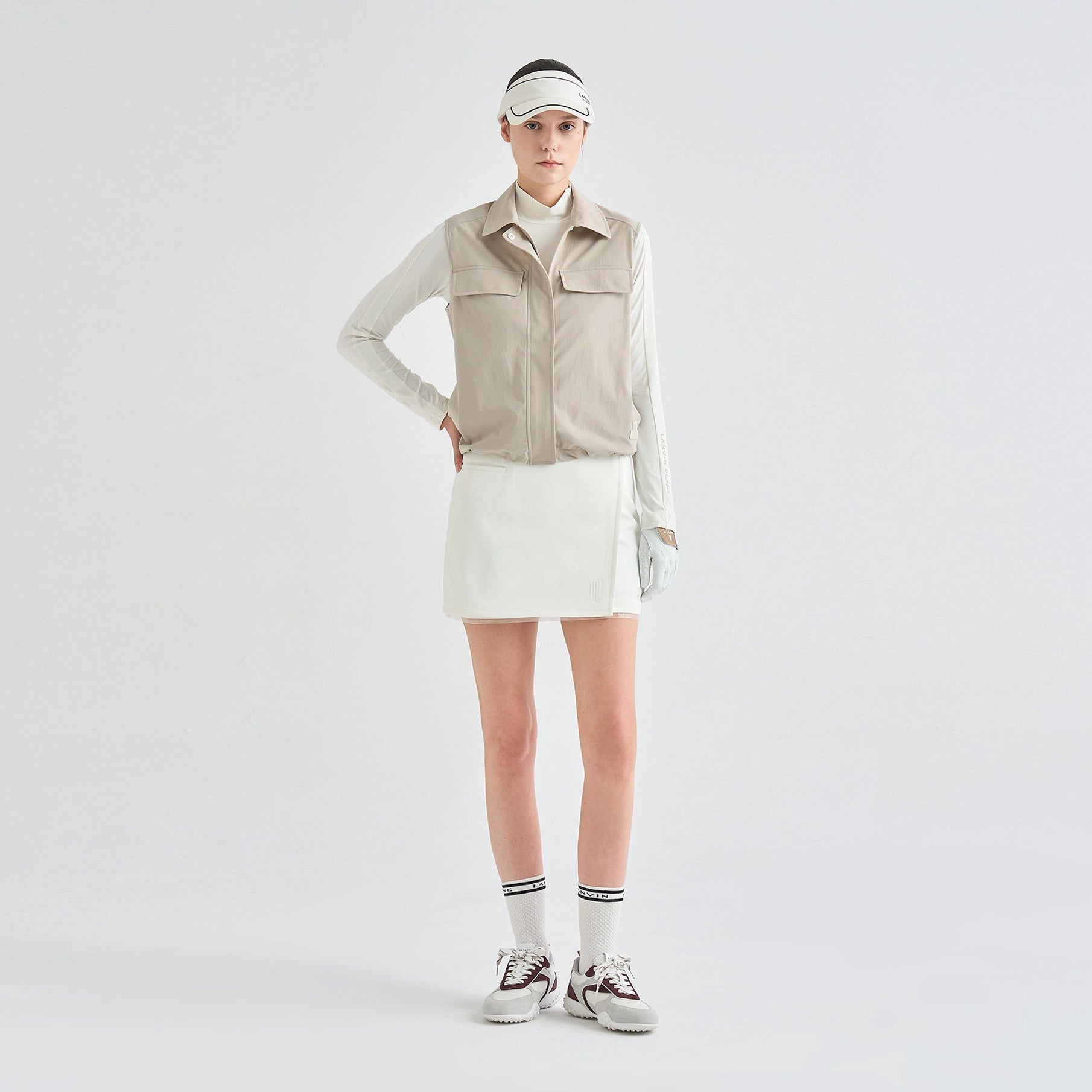 A model stands against a light gray studio background wearing a beige sleeveless zip-front vest over a white long-sleeve top, a white mini skirt, white socks, and white sneakers.