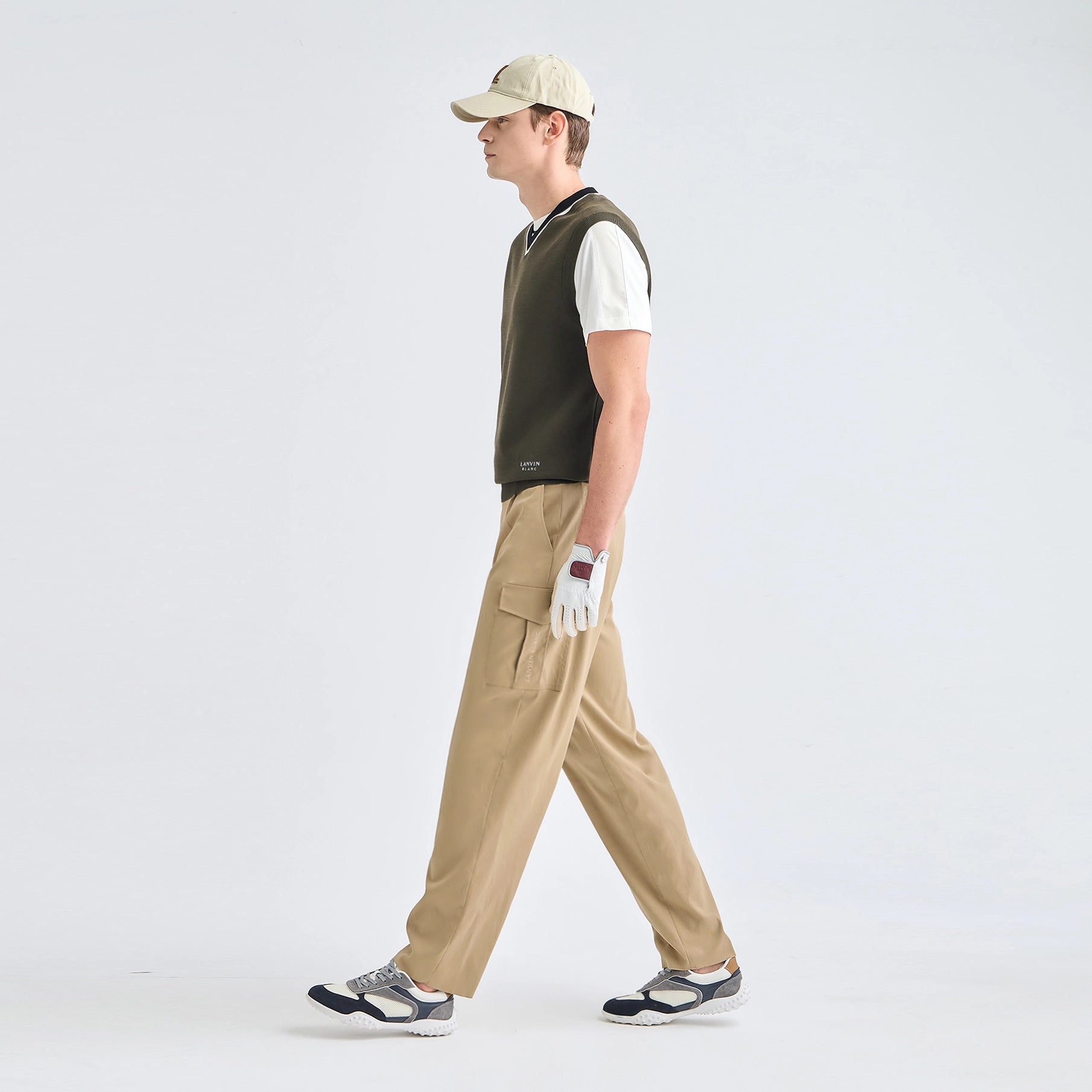 A man in profile wearing a beige baseball cap, a dark olive green knit vest over a white t-shirt, khaki cargo pants, and golf shoes, walking against a light gray studio background.