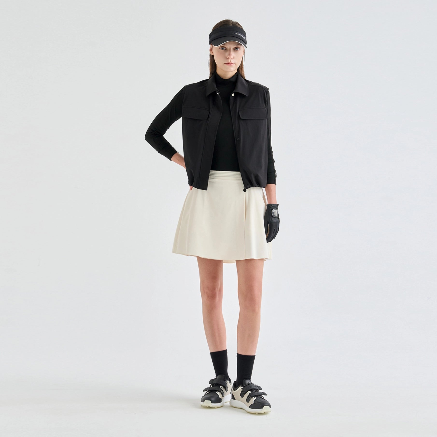 A model stands against a light gray studio background wearing a black long-sleeve collared jacket over a black top, a cream pleated mini skirt, black socks, black-and-white sneakers, and a black visor.