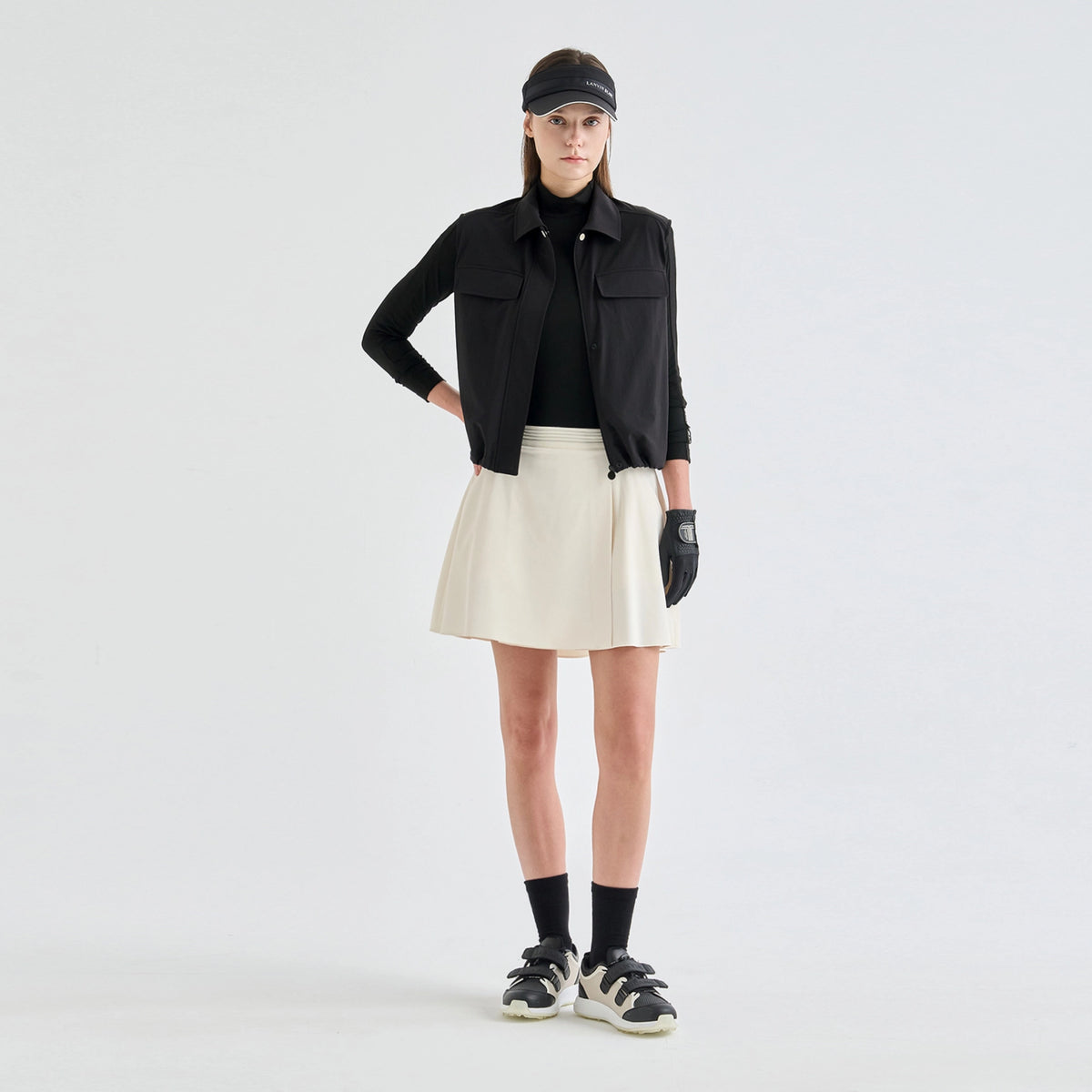 A model stands against a light gray studio background wearing a black long-sleeve collared jacket over a black top, a cream pleated mini skirt, black socks, black-and-white sneakers, and a black visor.