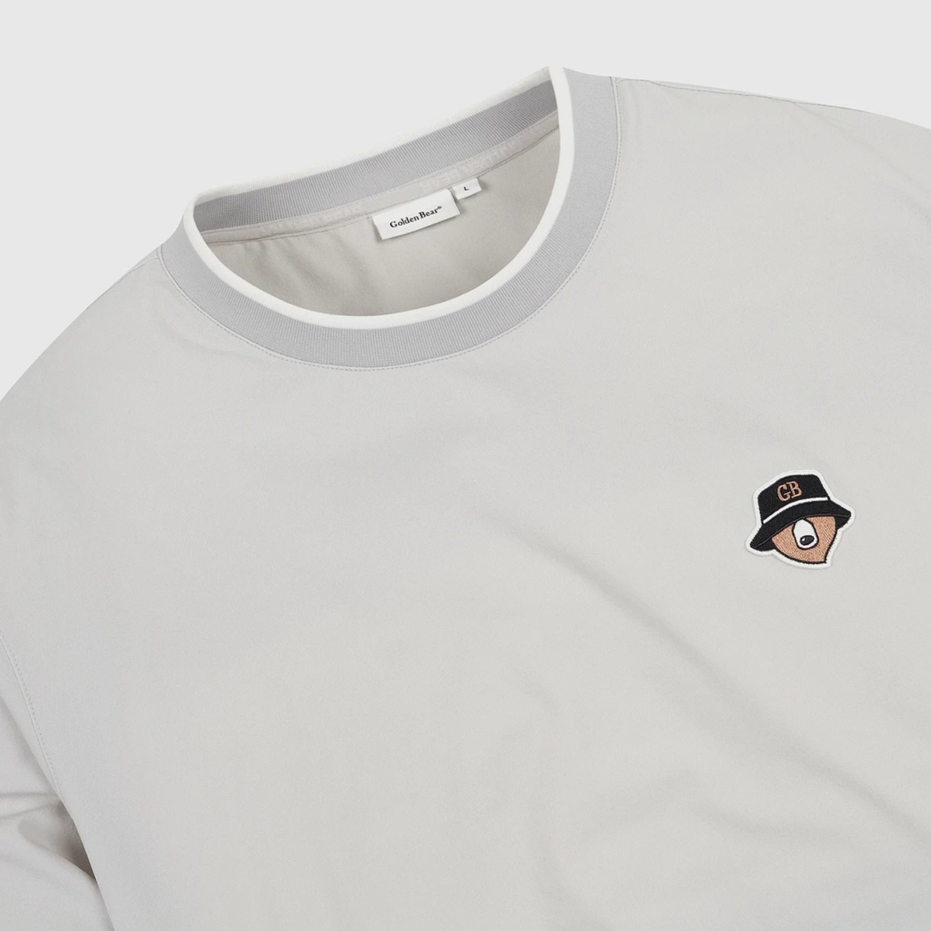 A close-up of a light gray crewneck sweatshirt with a gray ribbed collar, featuring a small embroidered cartoon eye wearing a black bucket hat with 'CB' on the chest.