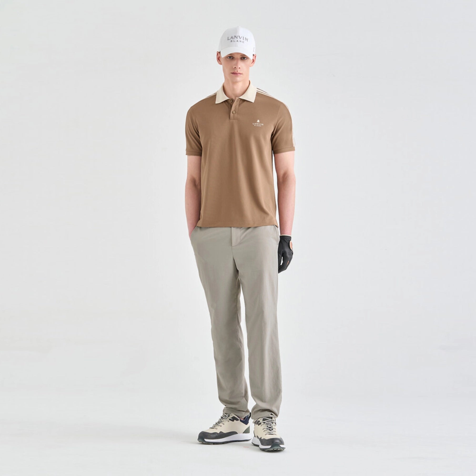 A man modeling a tan short-sleeve polo shirt with a cream collar, paired with light gray trousers, a white cap, a black glove, and golf shoes, standing against a light gray studio background.