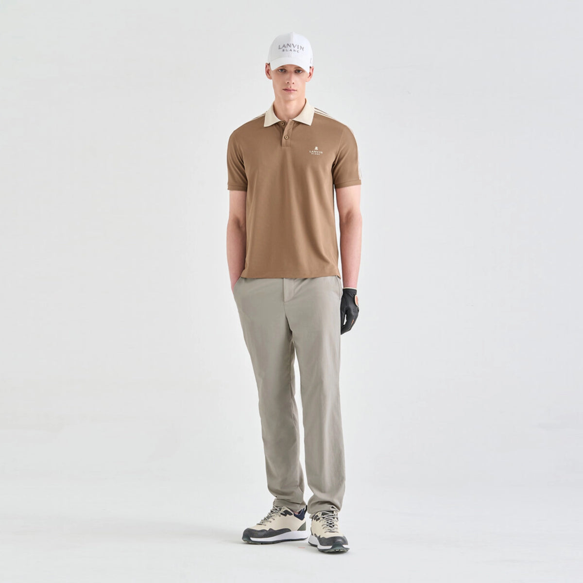 A man modeling a tan short-sleeve polo shirt with a cream collar, paired with light gray trousers, a white cap, a black glove, and golf shoes, standing against a light gray studio background.