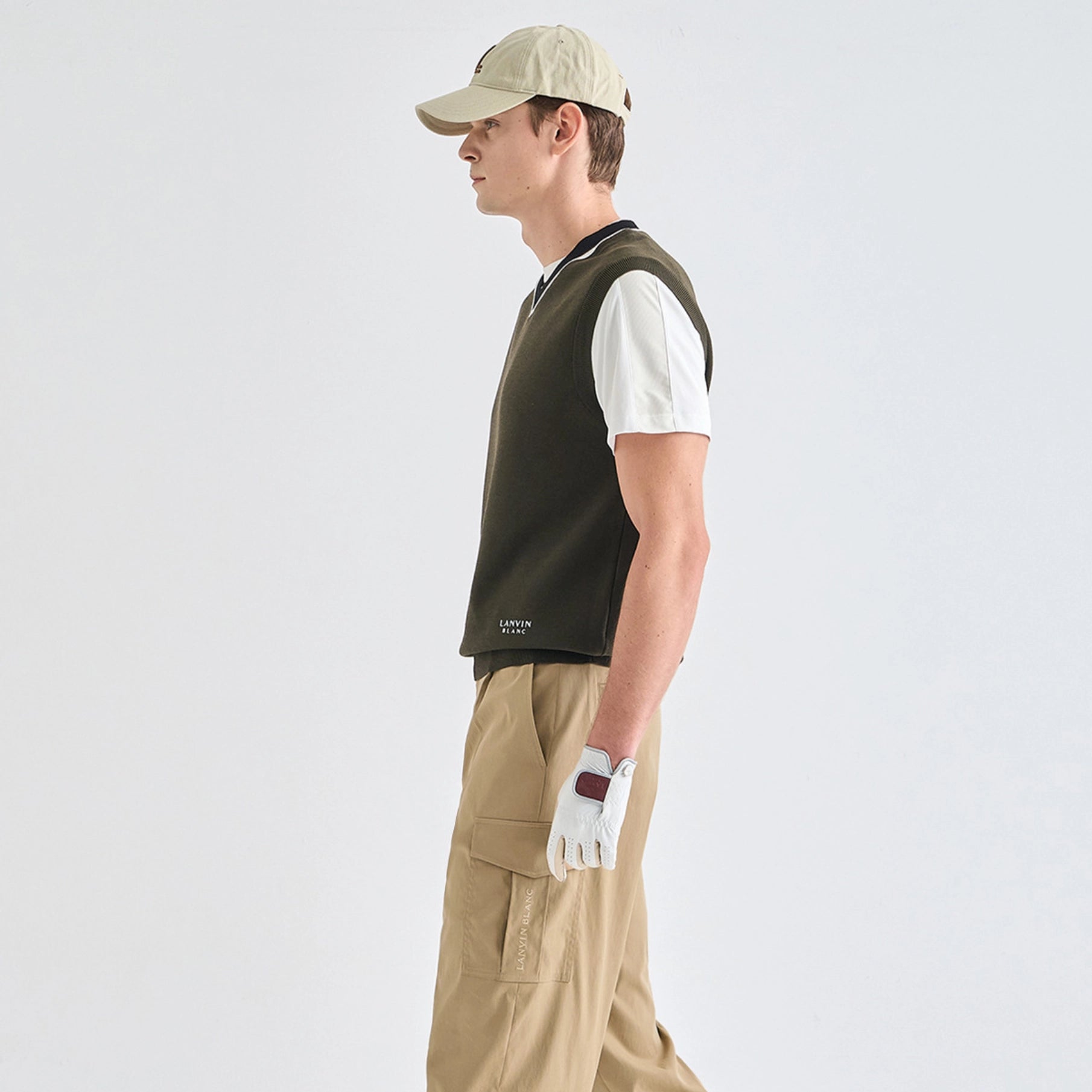 A man in profile wearing a dark olive green sleeveless polo vest over a white short-sleeve polo shirt, khaki cargo pants, a beige baseball cap, and a white golf glove, set against a plain light gray studio background.