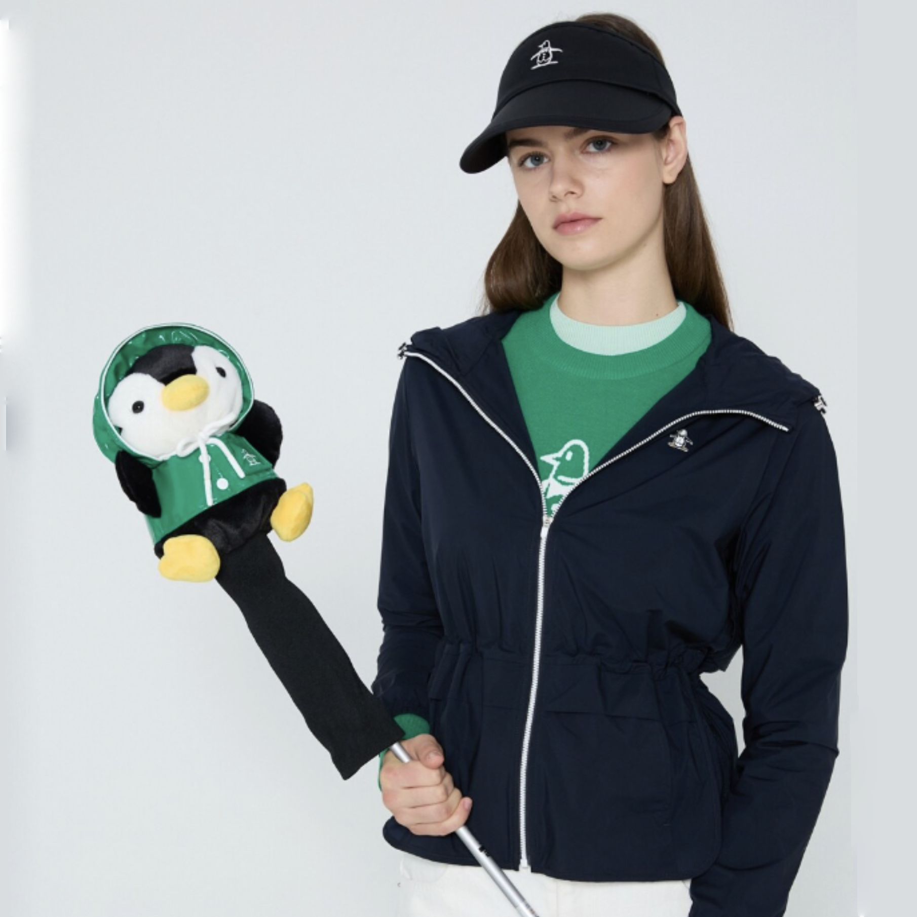 A woman wearing a navy zip-up jacket, green t-shirt, and black visor holds a golf club with a plush penguin headcover against a light gray studio background.