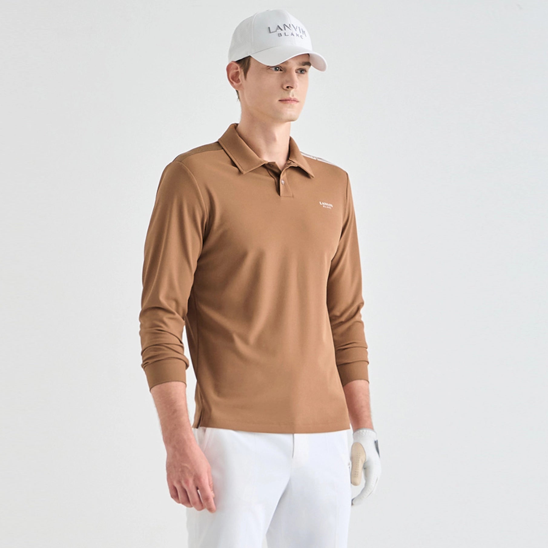 A man wearing a tan long-sleeve polo shirt, white pants, a white cap with 'LANEER' printed on it, and a white golf glove, standing against a light gray studio background.