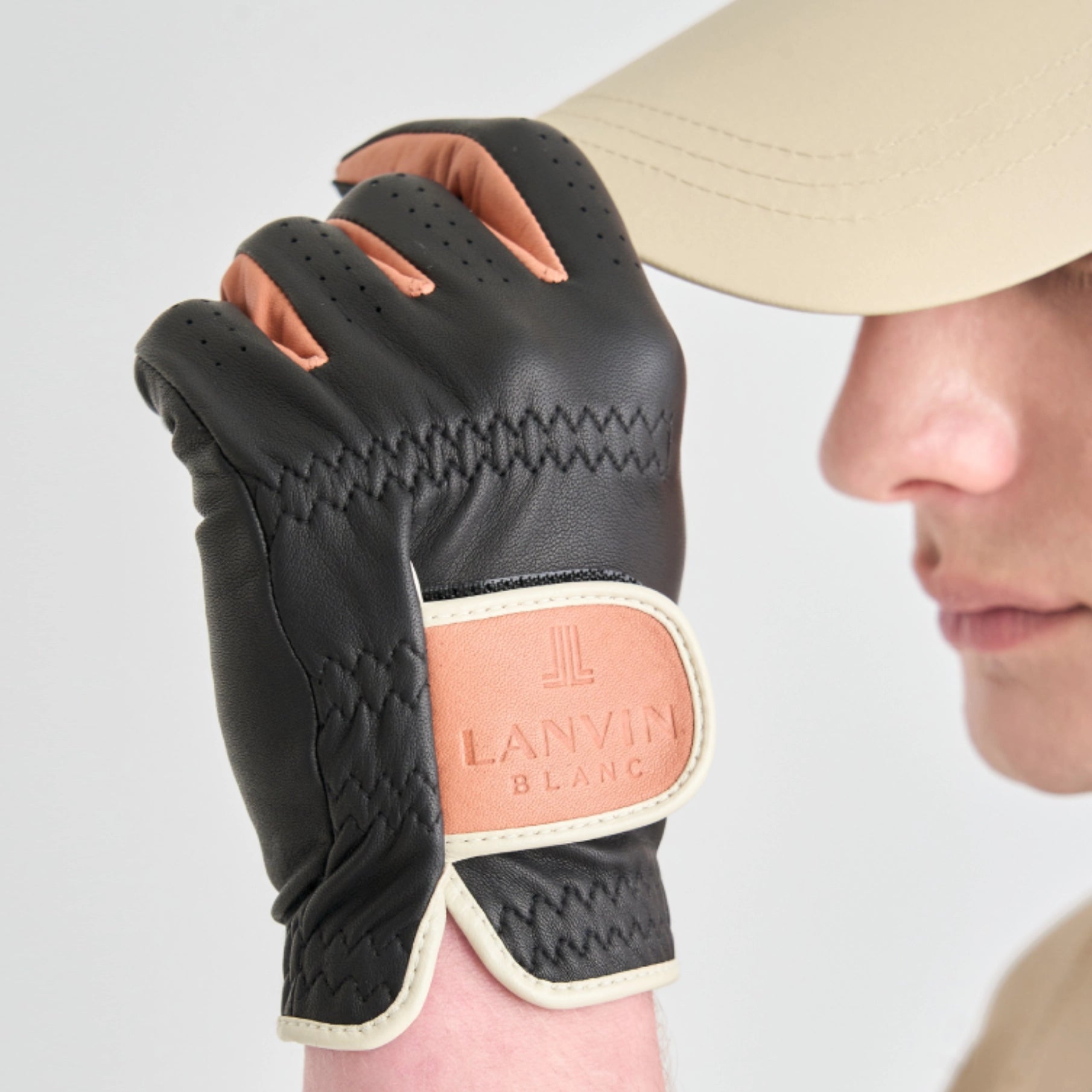 A close-up of a person wearing a black leather golf glove with a tan Lanvin Blanc patch on the back of the hand, holding a tan baseball cap against a light gray background.