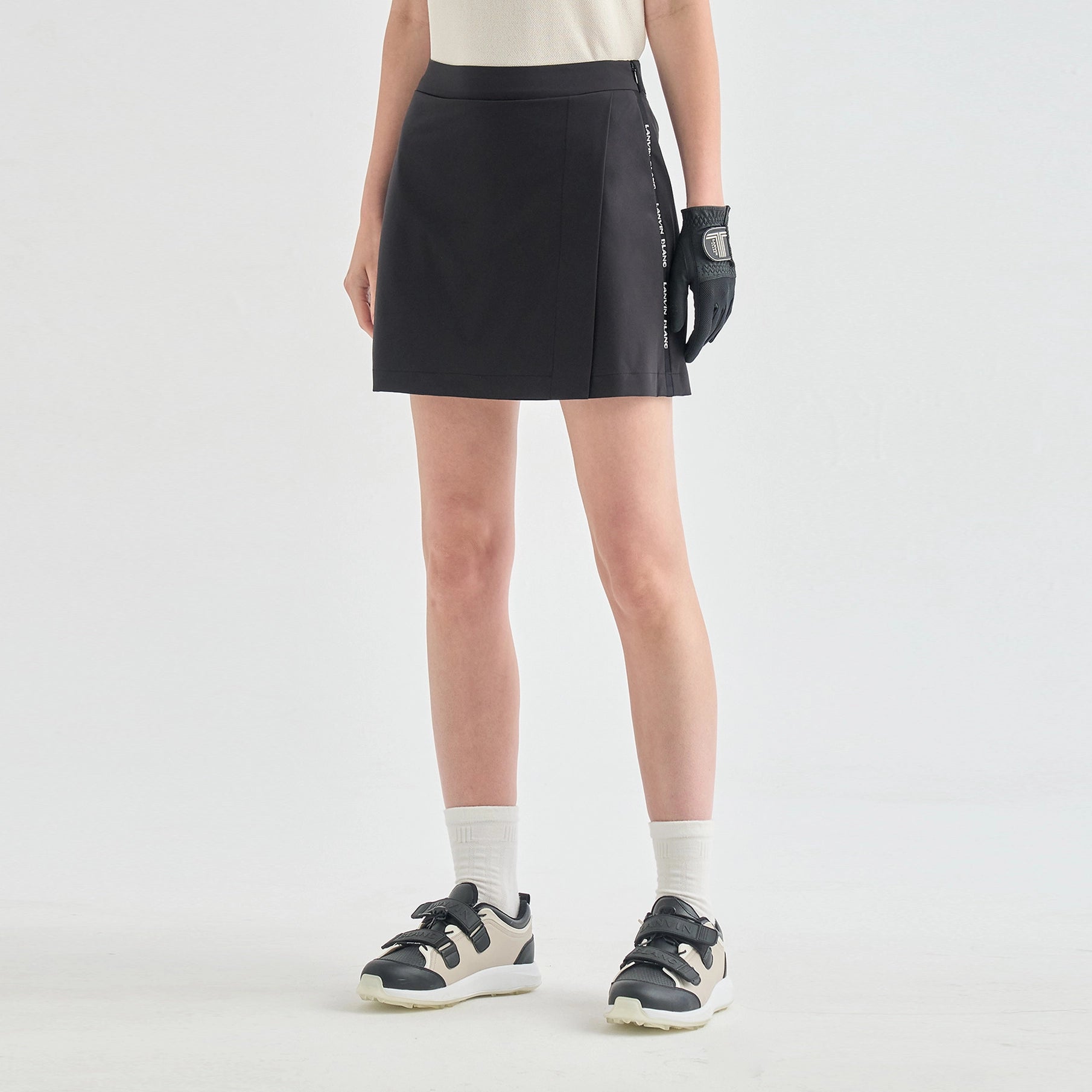 A model from the waist down wearing a black athletic mini skirt with white text on the side, paired with white socks, black and beige sneakers, and a black glove, against a light gray studio background.