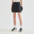 A model from the waist down wearing a black athletic mini skirt with white text on the side, paired with white socks, black and beige sneakers, and a black glove, against a light gray studio background.