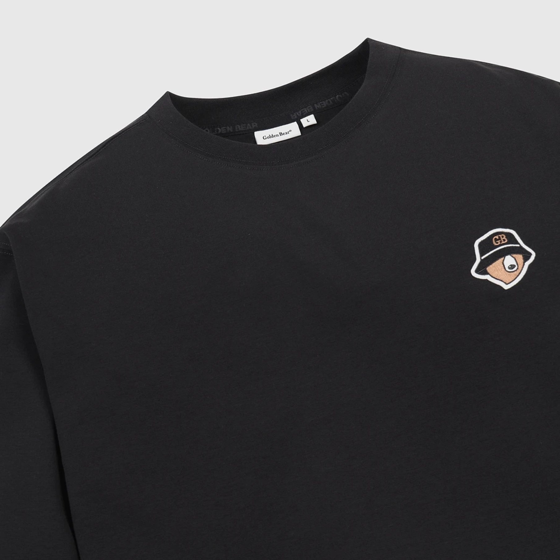 A close-up of a black crewneck sweatshirt featuring a small embroidered patch of a cartoon character wearing a bucket hat on the left chest. The brand name 'Golden Bear' is visible on the inner neck label.