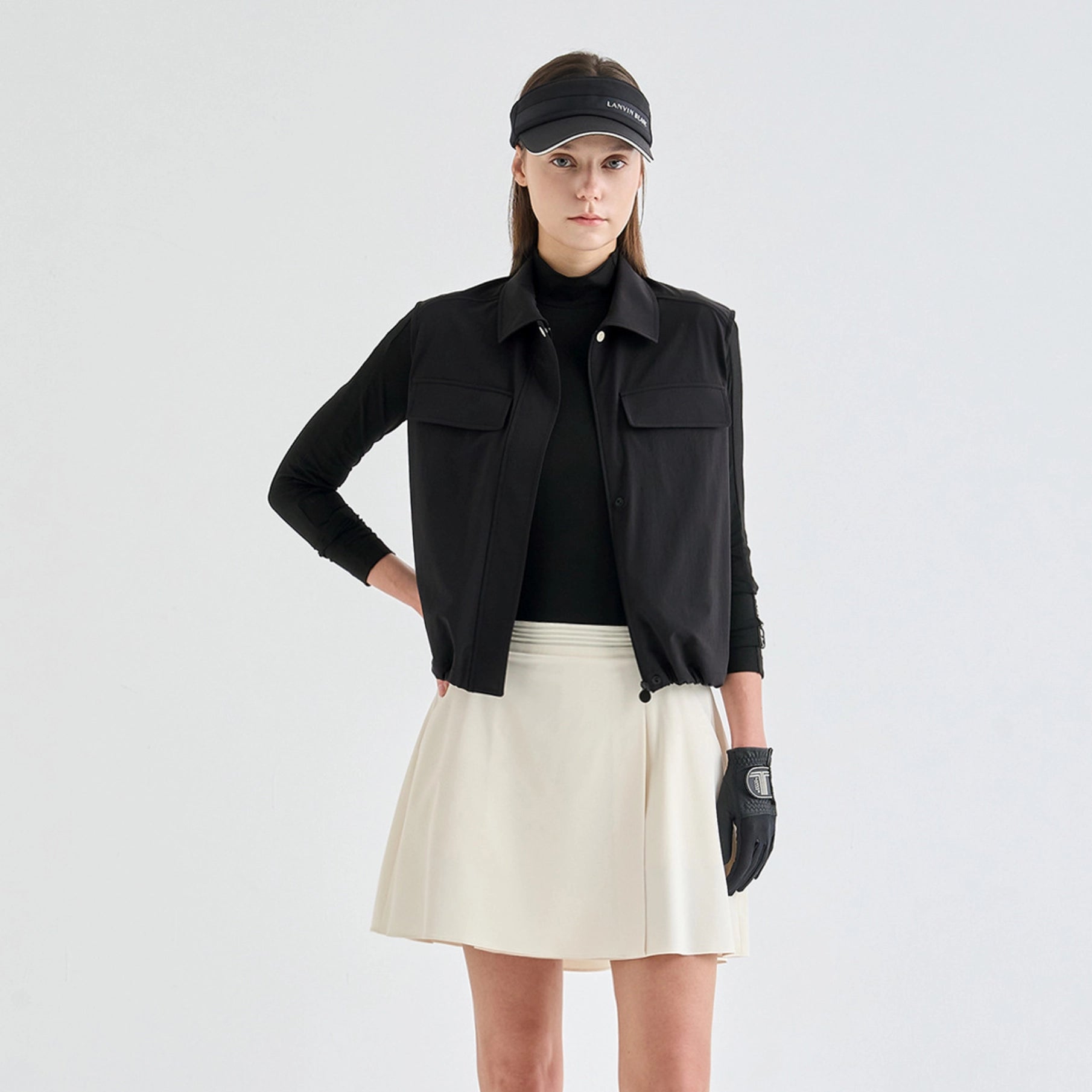 A model stands against a light gray studio background wearing a black sleeveless collared jacket over a black turtleneck, a light cream pleated mini skirt, a black visor, and a black glove.