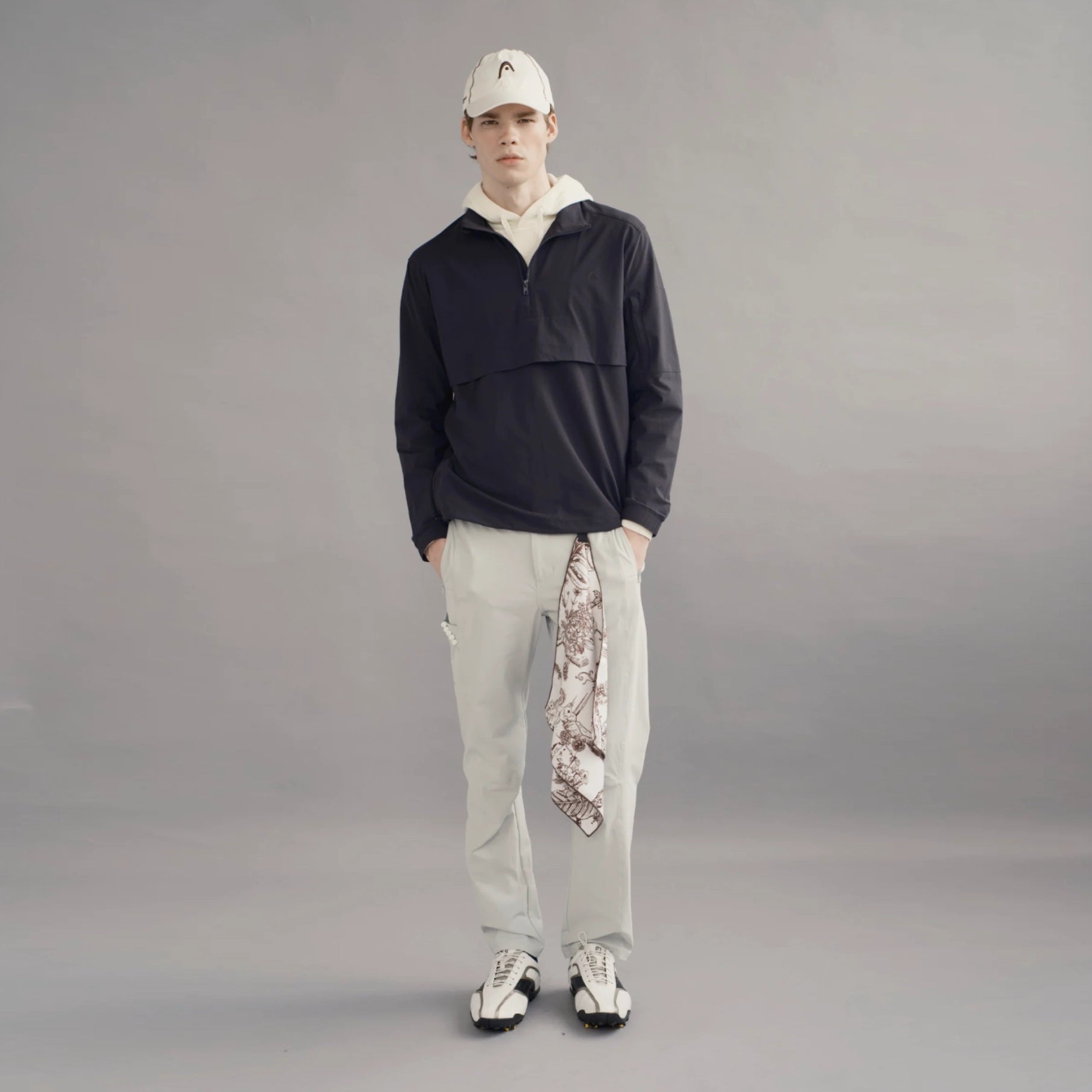 A male model stands against a neutral gray studio background wearing a dark navy half-zip pullover, light gray sweatpants, a white baseball cap, and white sneakers. A patterned scarf is draped over one leg.