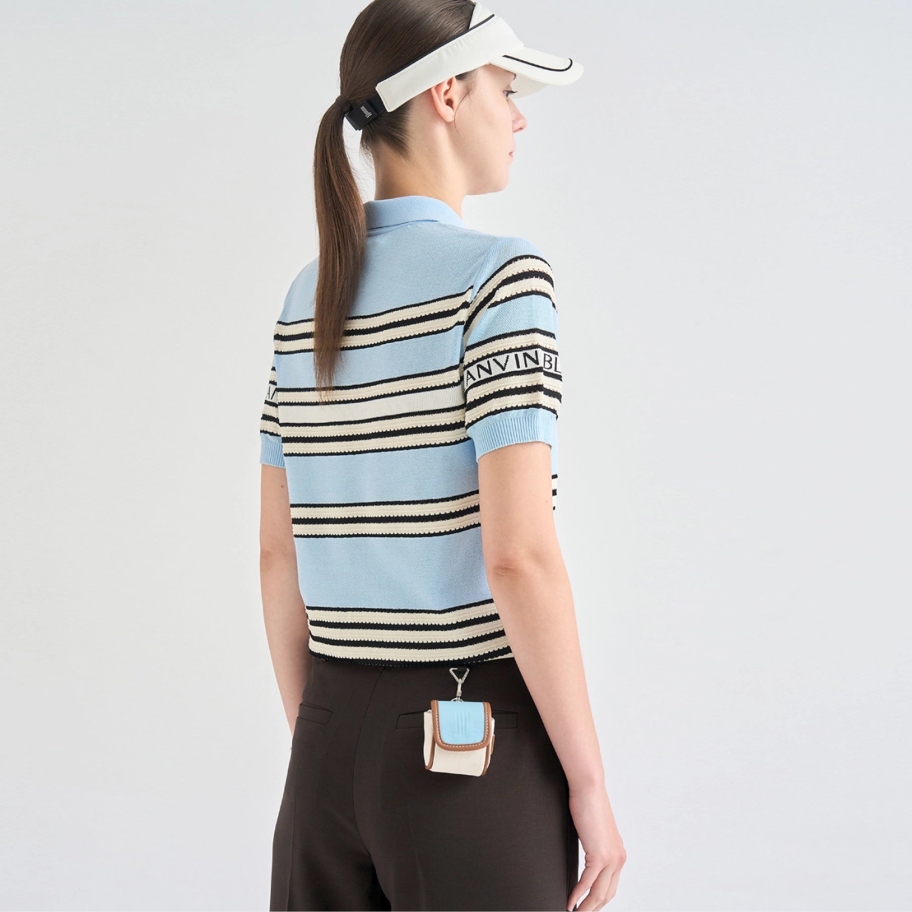 A woman shown from the back wearing a light blue short-sleeve knit polo shirt with black and beige horizontal stripes, a white visor, and dark brown pants. The brand name 'MARVIN B.