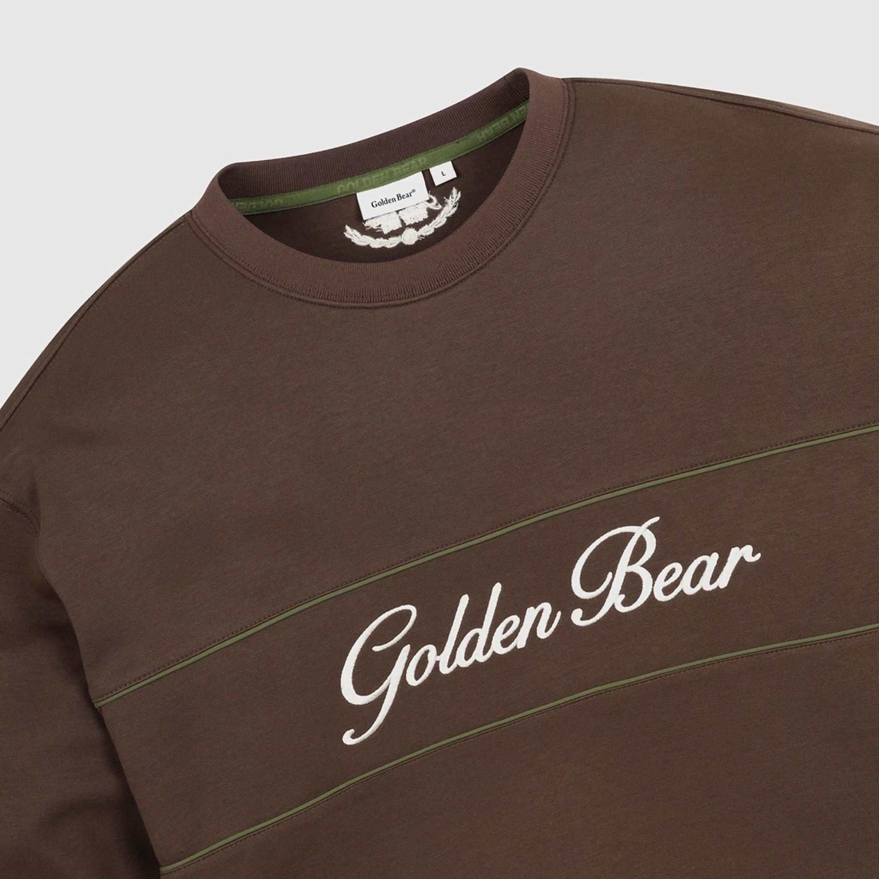 A close-up of a brown crewneck sweatshirt featuring 'Golden Bear' embroidered in white script across the chest, with thin green accent stripes and a visible inner neck label.