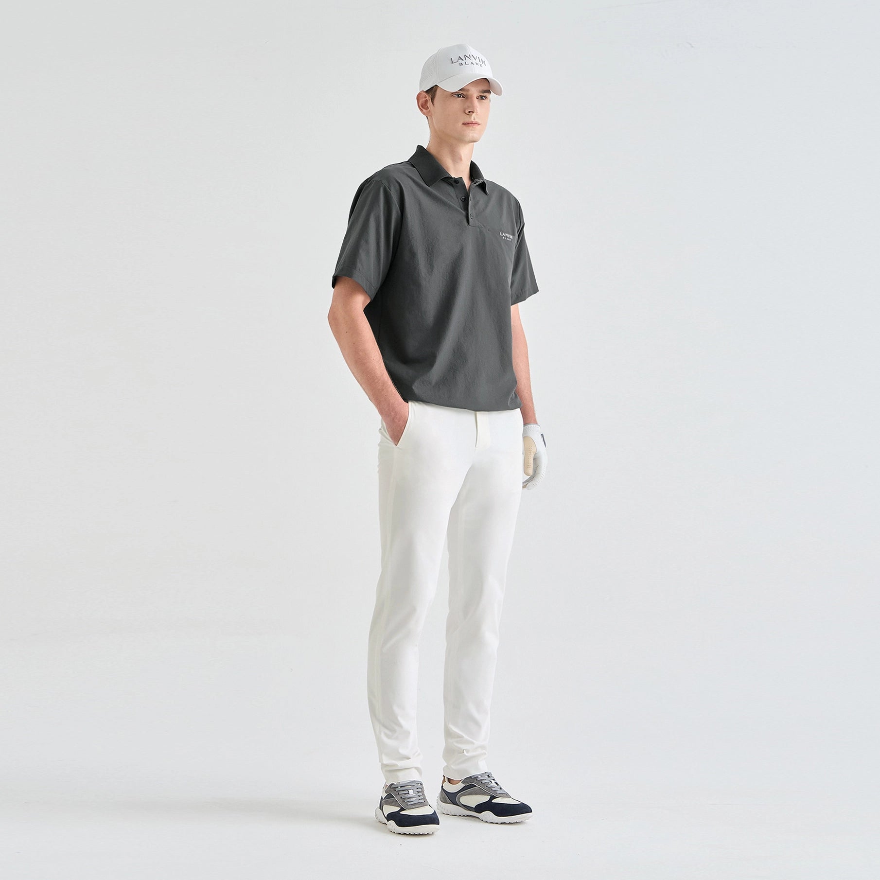 A man modeling a dark gray short-sleeve polo shirt, white golf pants, a white cap, and white golf shoes against a plain light gray studio background.