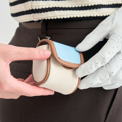 A close-up of a golf accessory pouch being held by hands and a white perforated golf glove. The pouch has a cream-colored textured body, brown leather trim, and a light blue flap.