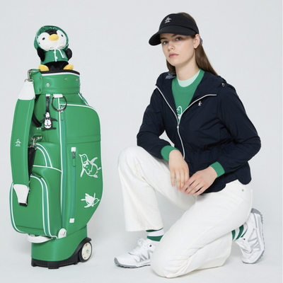 A woman kneels beside a bright green golf bag with a penguin plush on top, wearing a navy zip jacket, green shirt, white pants, and white sneakers against a light gray studio background.