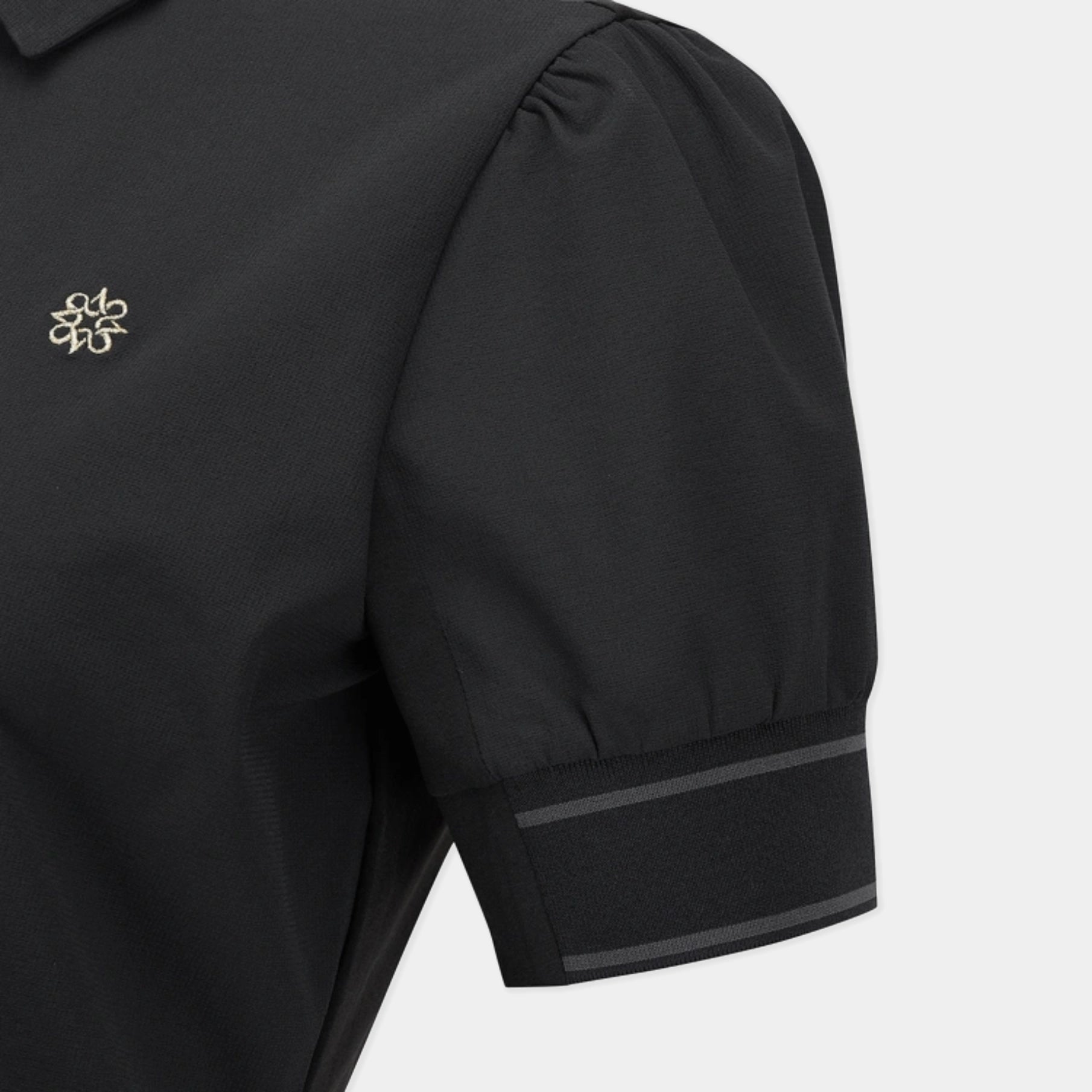 Close-up of a black short-sleeve blouse with a small gold embroidered logo on the chest and a double-stripe detail on the sleeve cuff, shown against a light gray background.