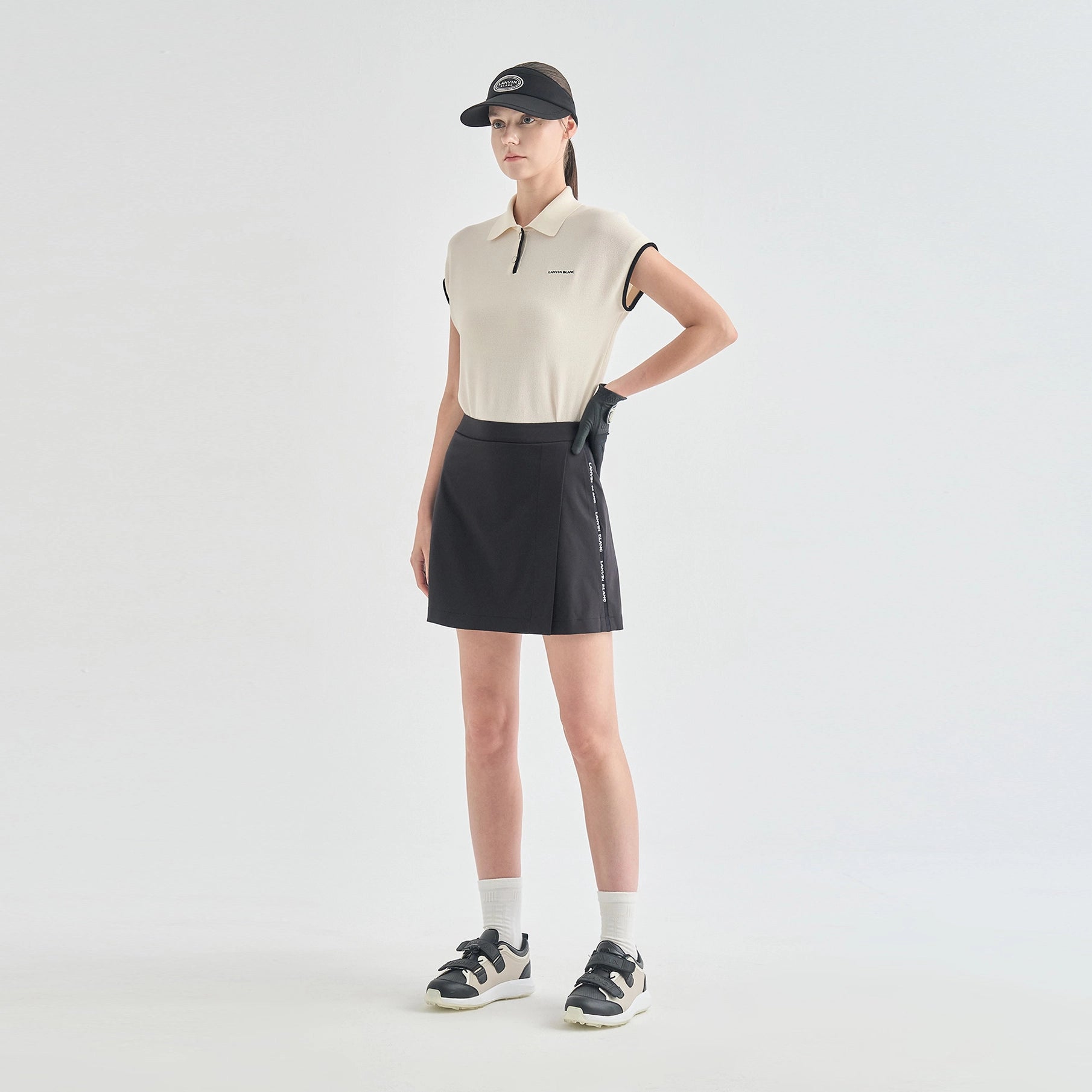 A model stands in a studio wearing a sleeveless cream polo shirt, a black athletic mini skirt with white side lettering, a black cap, white socks, and black-and-white sneakers.