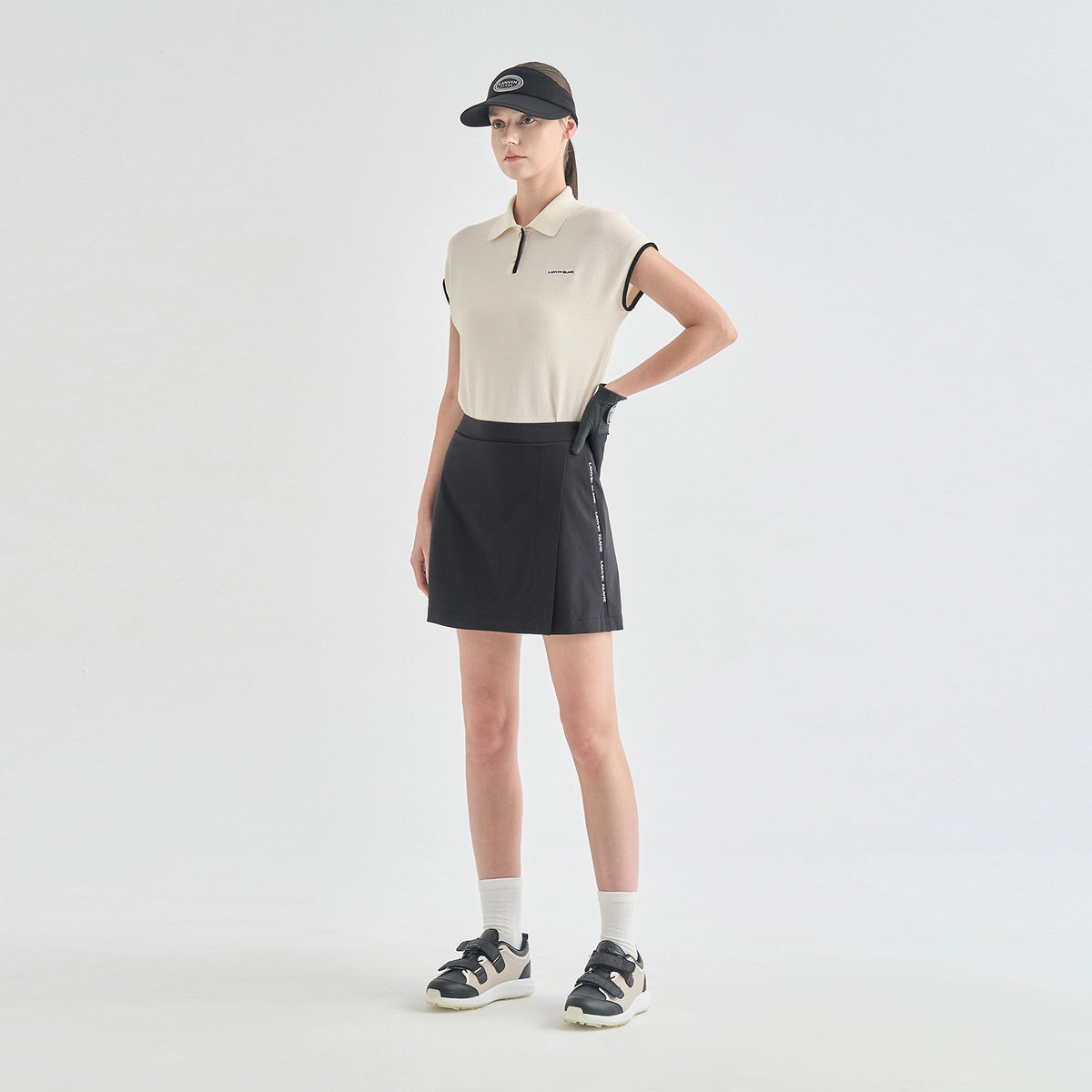 A model stands in a studio wearing a sleeveless cream polo shirt, a black athletic mini skirt with white side lettering, a black cap, white socks, and black-and-white sneakers.