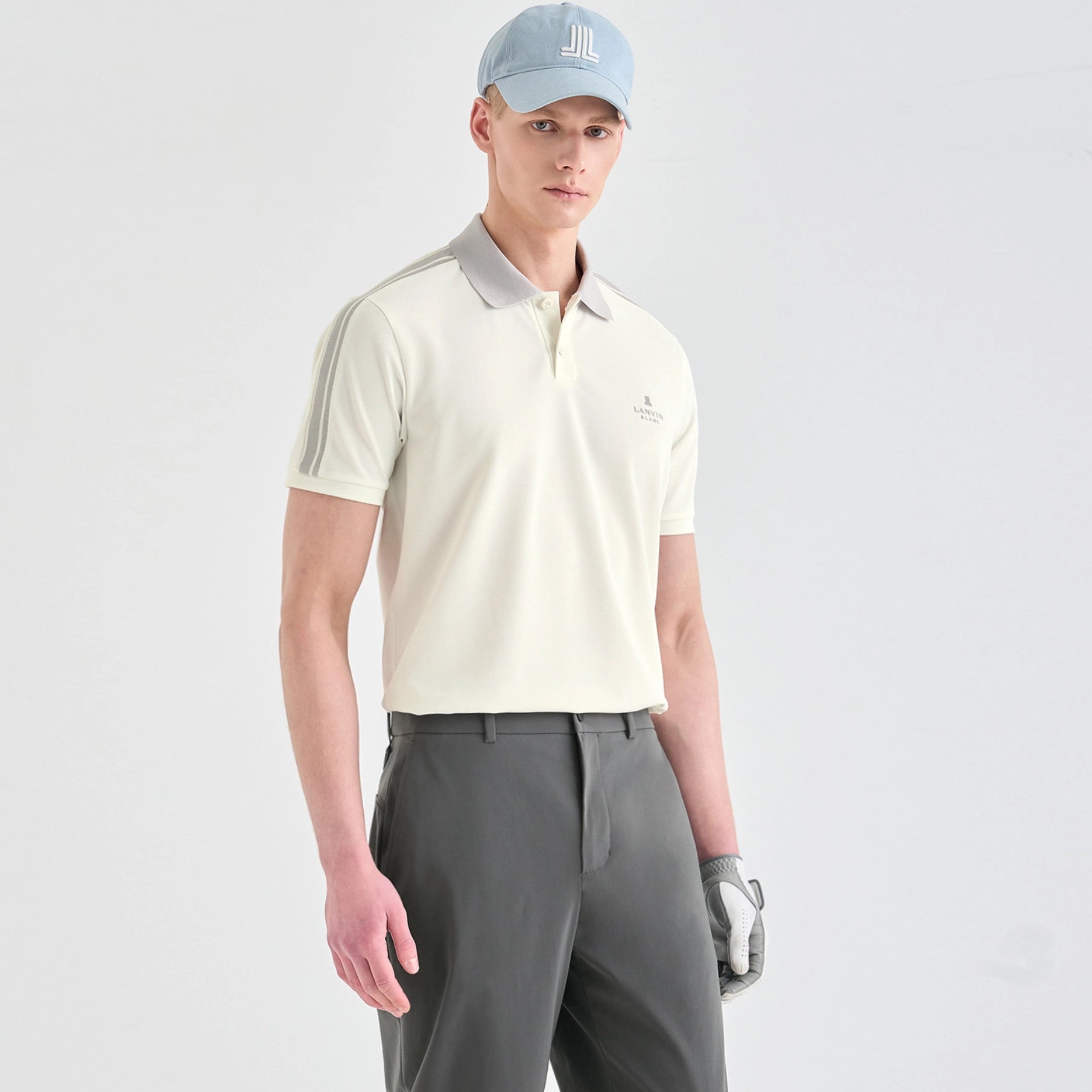 A male model wearing a light cream-colored short-sleeve polo shirt with gray accents on the collar and shoulders, paired with gray trousers, a light blue cap, and a golf glove, set against a plain light gray studio background.