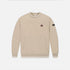 A beige crewneck sweatshirt with a small embroidered character patch on the left chest and a black brand patch on the right sleeve, displayed on a clean white background.