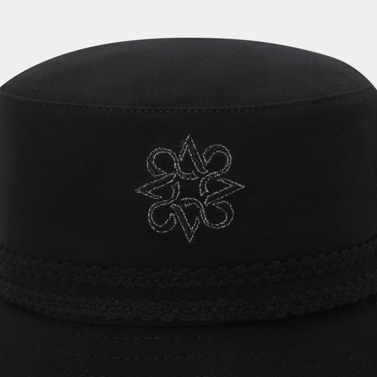 A close-up of a black fabric cap with a braided band and a white embroidered geometric logo on the front, set against a light gray background.