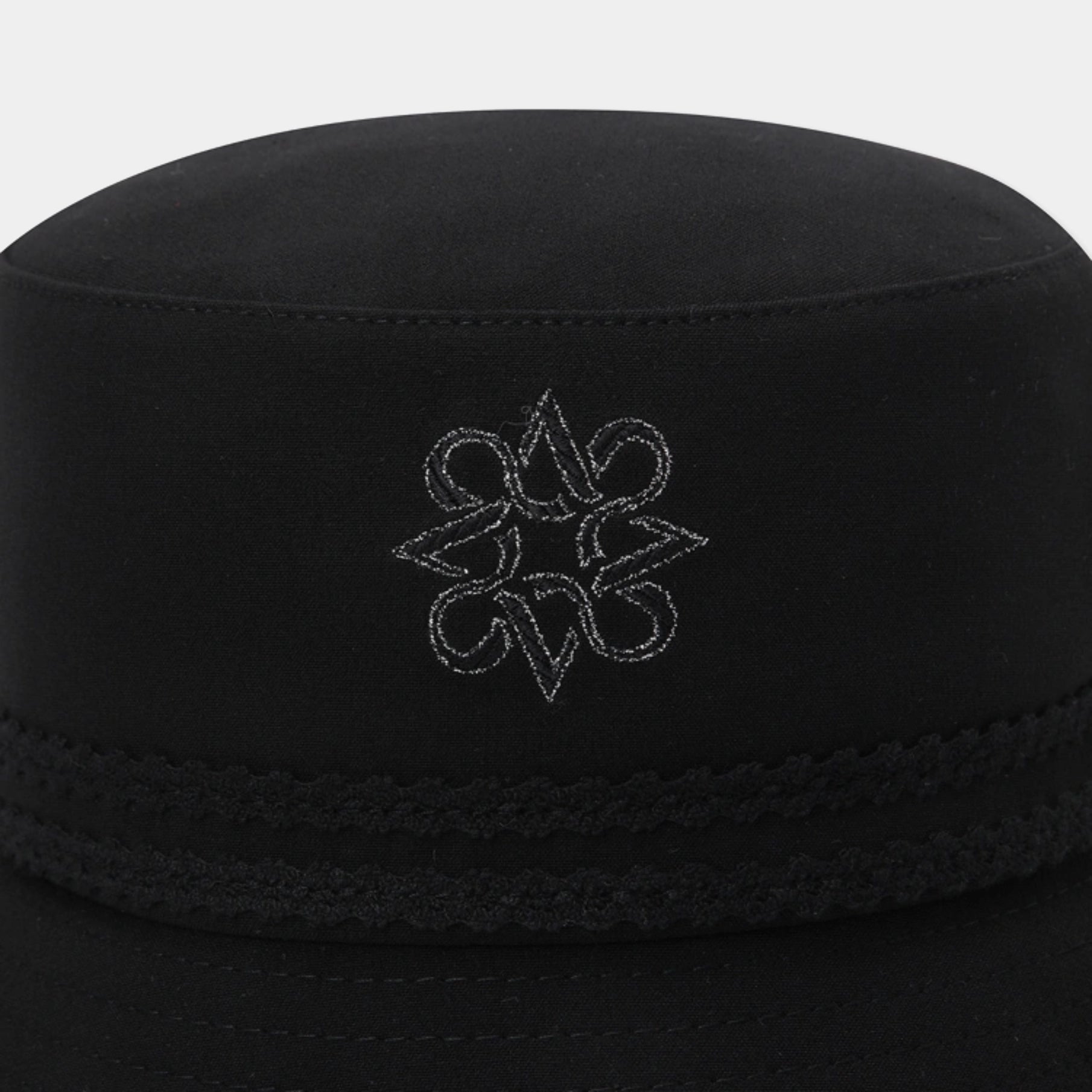 A close-up of a black fabric cap with a braided band and a white embroidered geometric logo on the front, set against a light gray background.