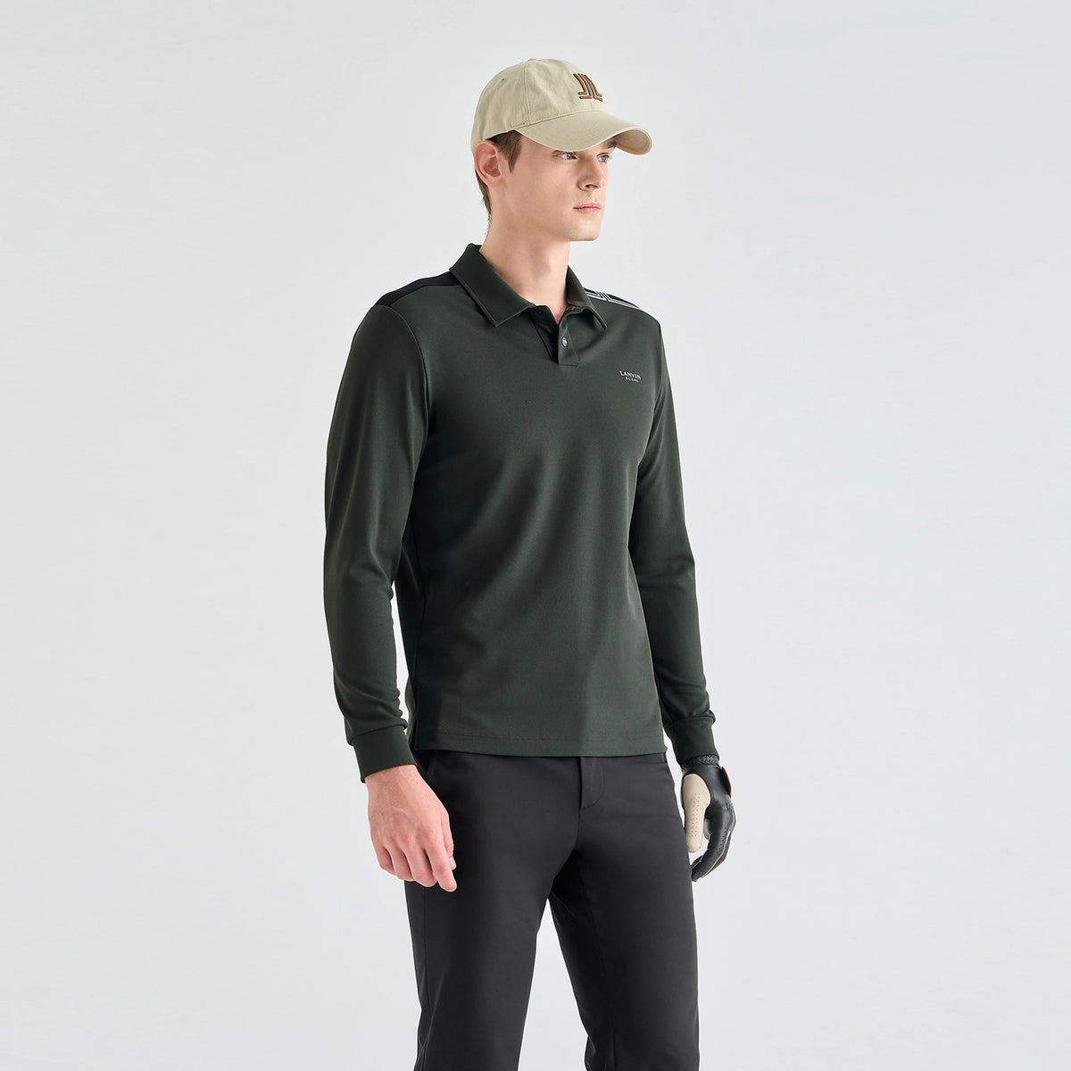 A man wearing a dark forest green long-sleeve polo shirt, black pants, a beige baseball cap, and a black golf glove, standing against a light gray studio background.