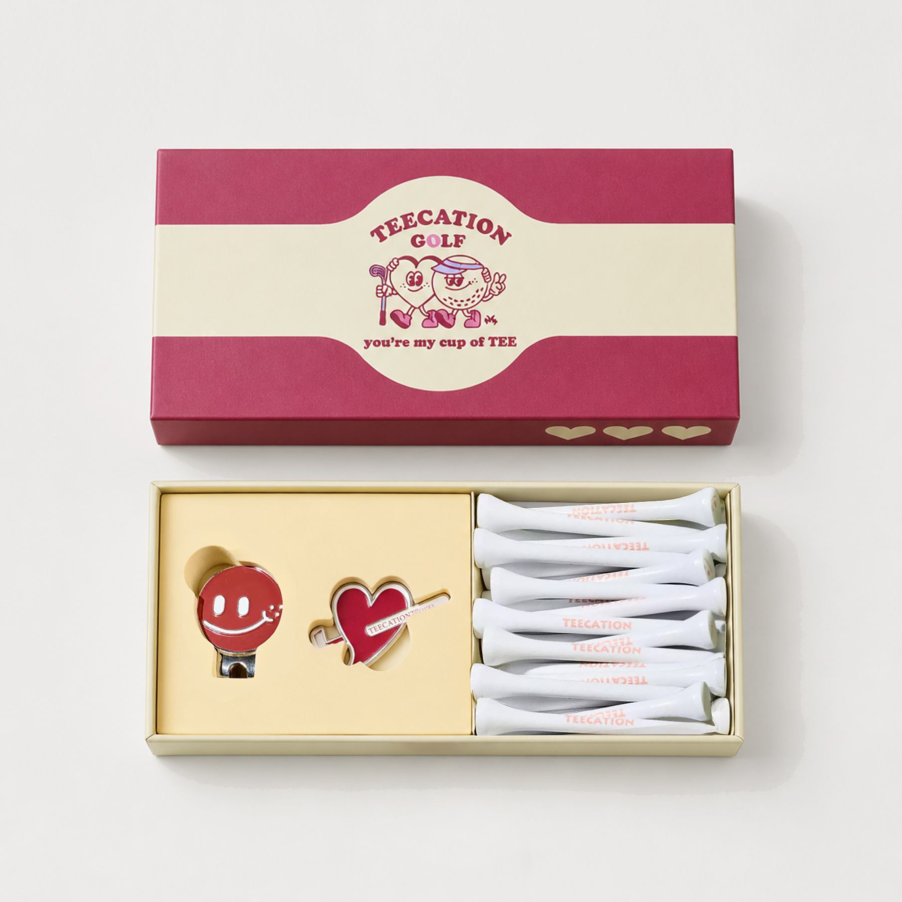 A boxed gift set on a light gray background, featuring a magenta and cream presentation box above an open tray containing two enamel pins and several white wrapped items. The packaging and pins show the visible brand name Teecation G.