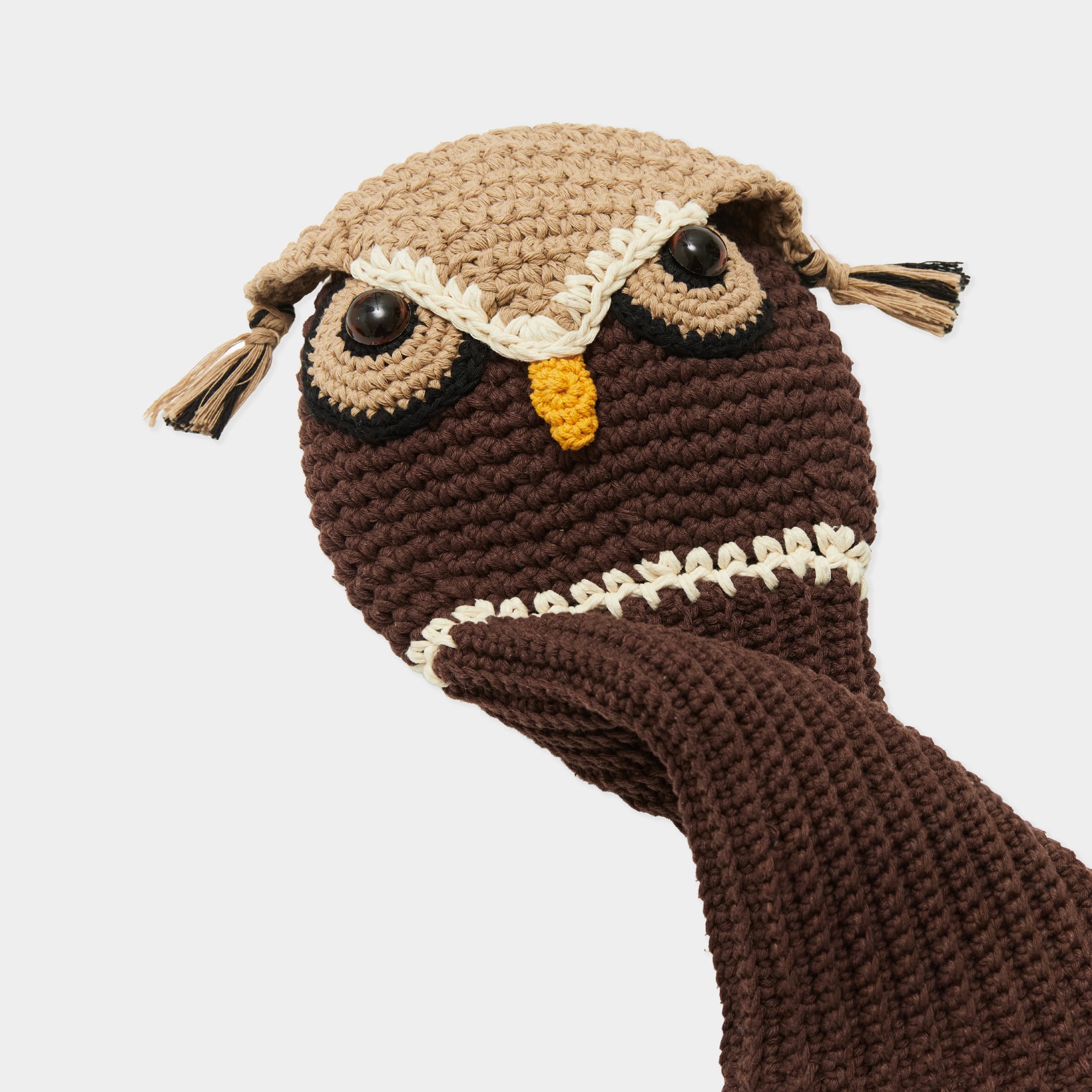 A close-up of a crocheted owl puppet head and hand puppet body in brown and beige, with black eyes, a yellow beak, and cream stitched details, shown against a light gray background.