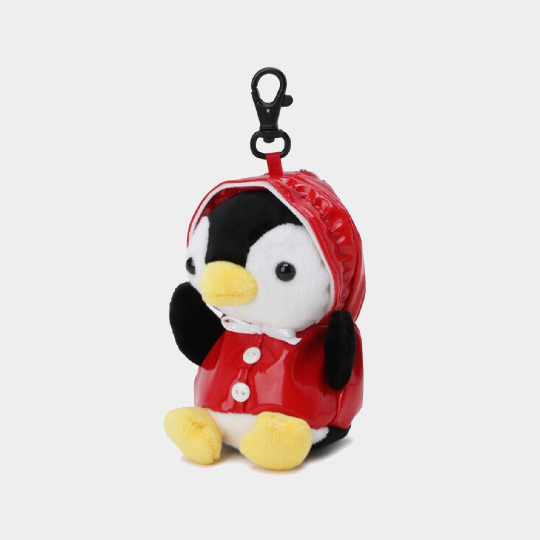 A plush penguin keychain or bag charm wearing a shiny red hooded raincoat with white buttons, black wings, a yellow beak and feet, and a black clasp on top, shown on a light gray background.