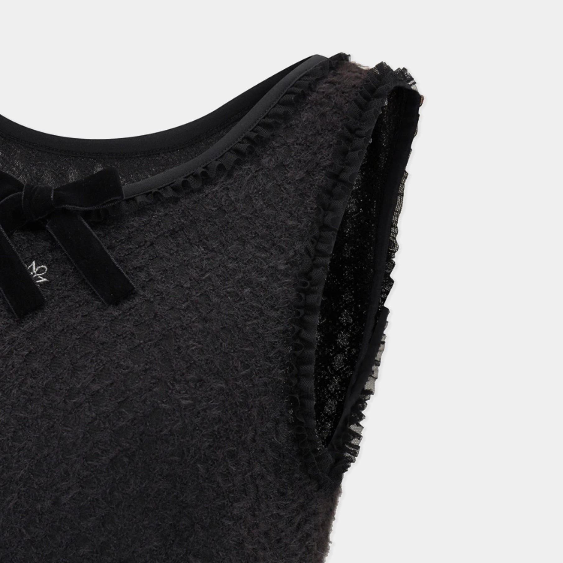 Close-up of a black textured sleeveless top with a ruffled neckline, black velvet bow, and lace trim on a light gray background.