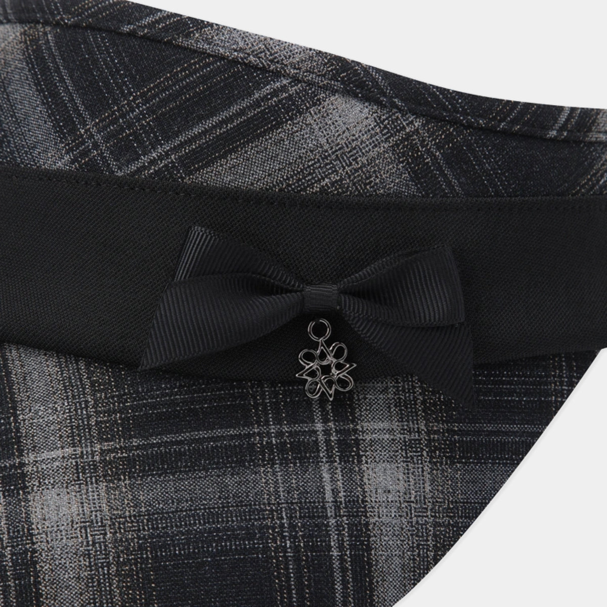 Close-up of a black ribbon bow with a small silver flower charm, attached to a black waistband with a gray and black plaid fabric.