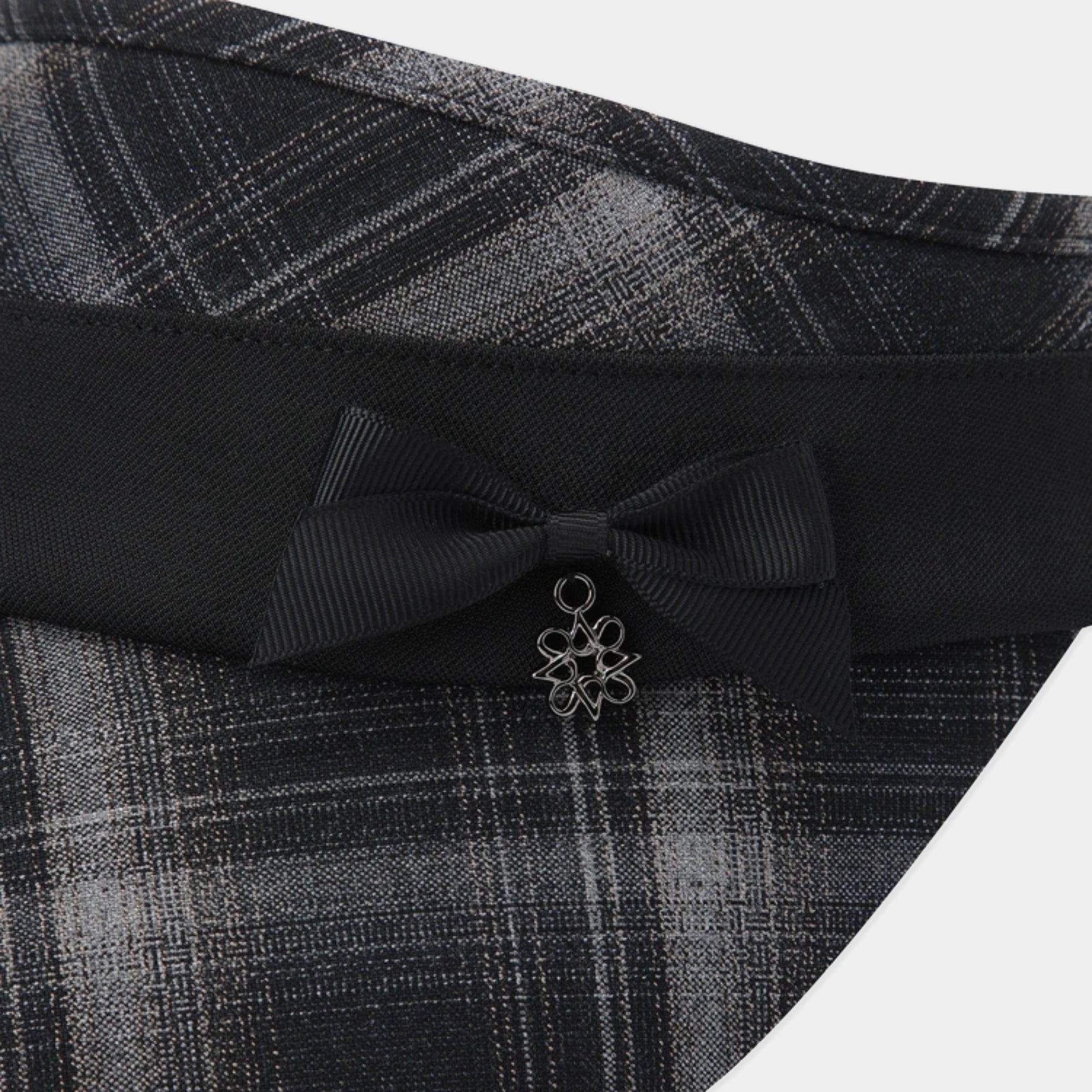 Close-up of a black ribbon bow with a small silver flower charm, attached to a black waistband with a gray and black plaid fabric.