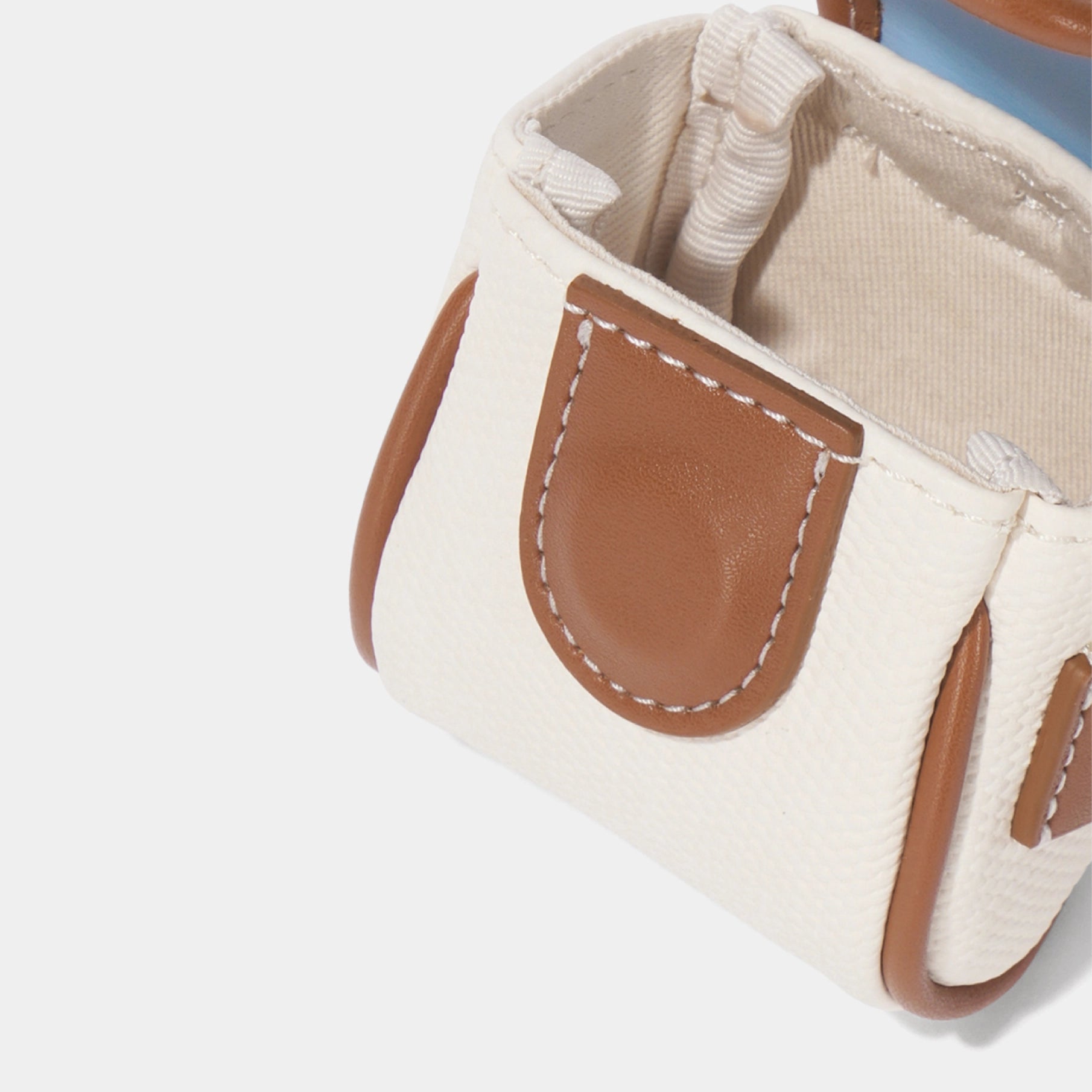 Close-up of a small structured handbag in off-white canvas with tan leather trim and white contrast stitching, shown on a light gray background.