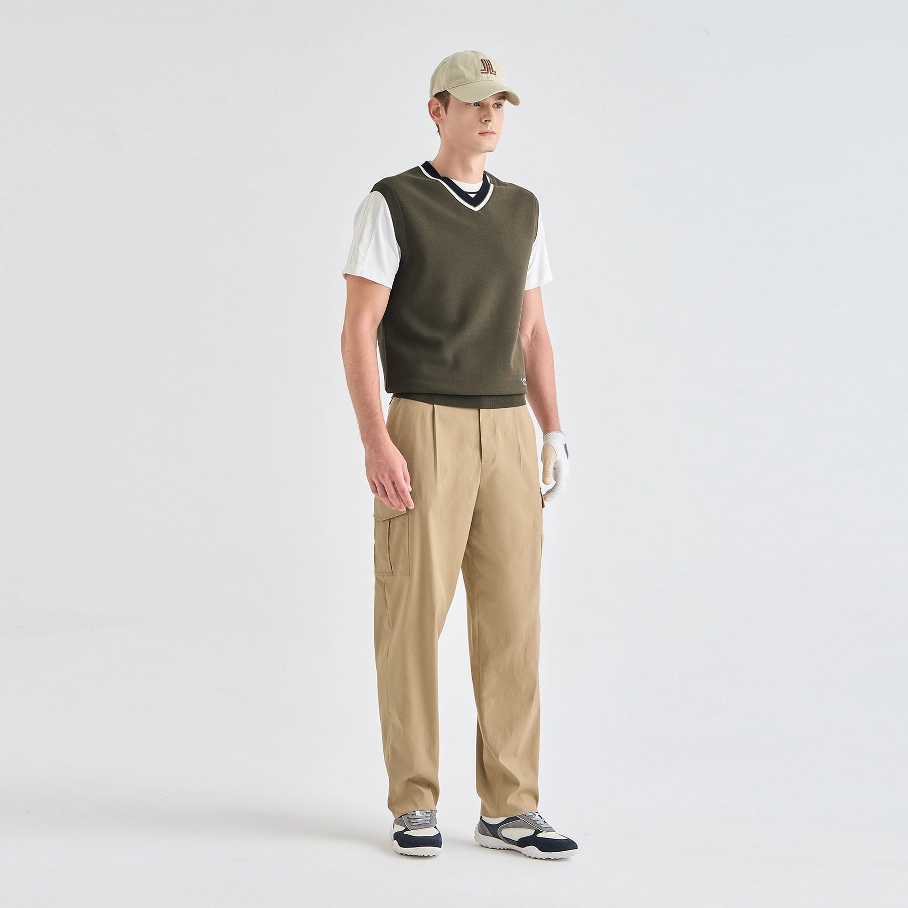 A man modeling a golf outfit consisting of a dark olive green knit vest over a white short-sleeve shirt, khaki pants, a beige baseball cap, and golf shoes, photographed against a seamless light gray studio background.