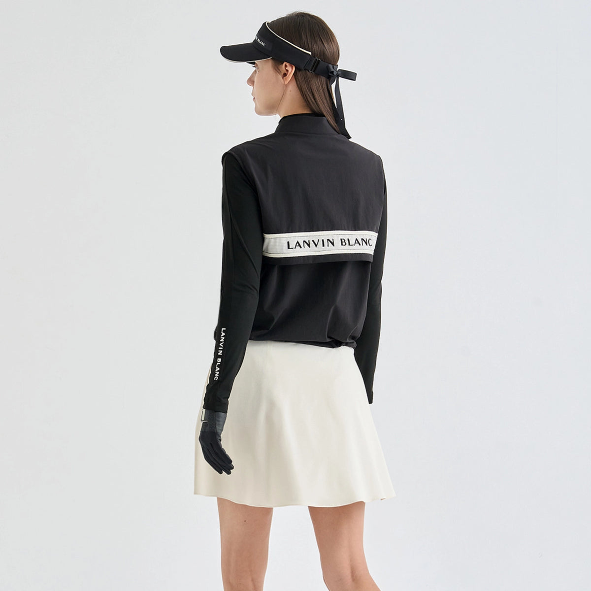A model shown from the back wearing a black athletic jacket with a white horizontal stripe that reads 'LANVIN BLANC', a white pleated skirt, black gloves, and a black visor against a light gray studio background.