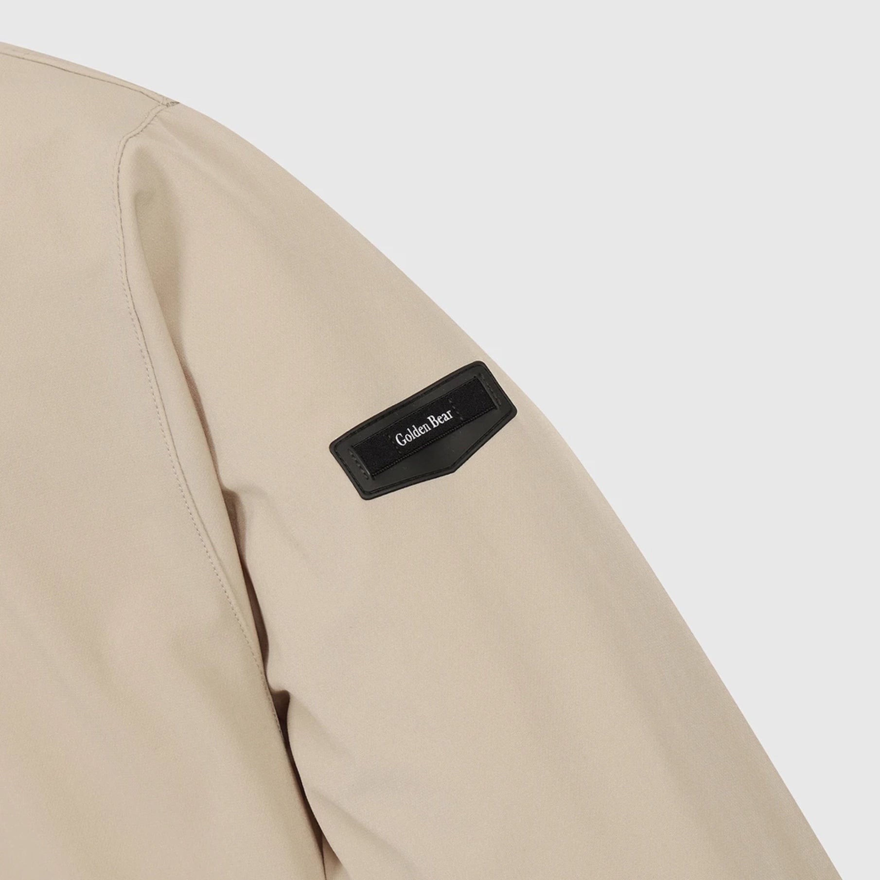 Close-up of a beige jacket sleeve featuring a black rectangular patch with the brand name 'Gola Gear' in white text, set against a light gray background.