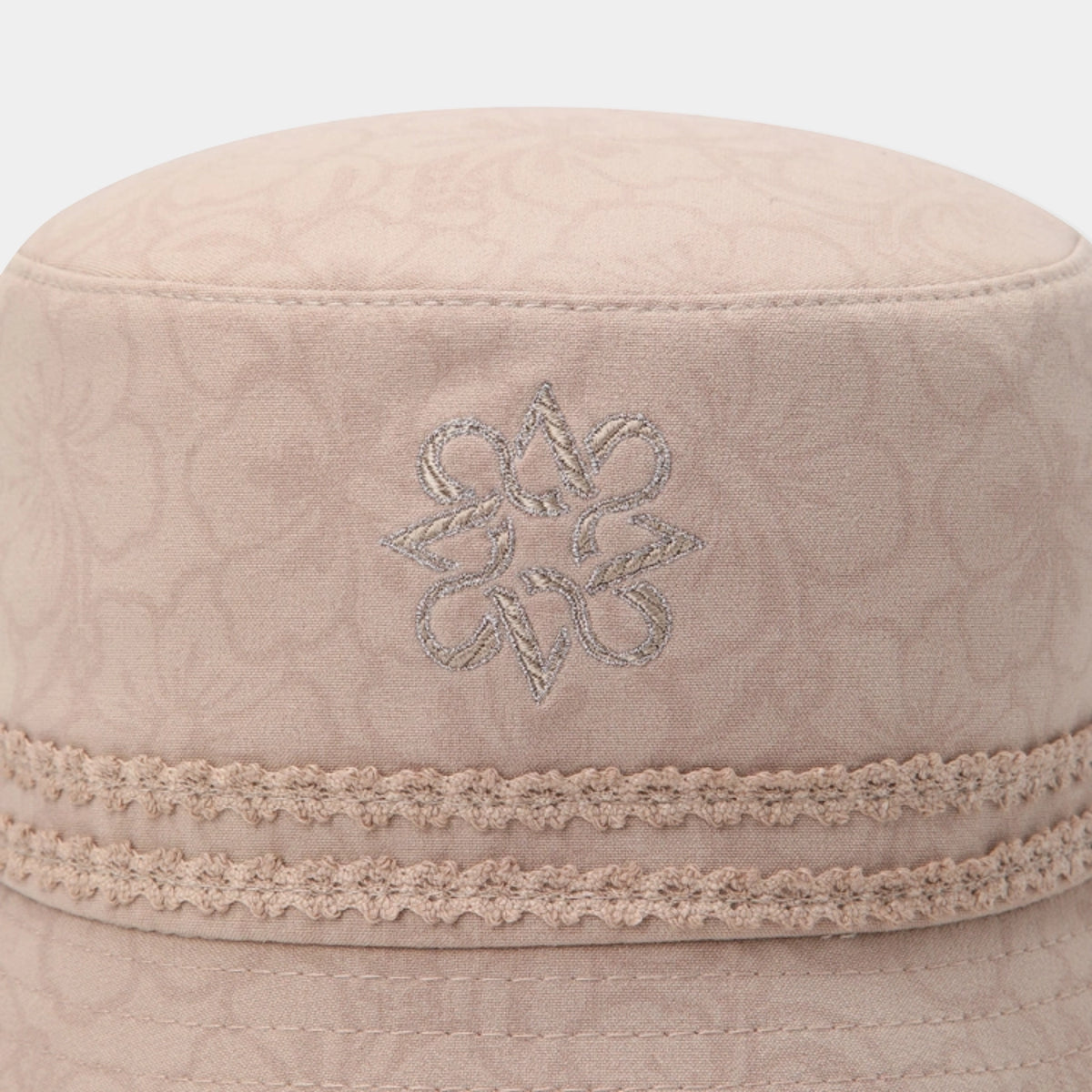 A close-up of a light beige structured cap featuring a subtle embossed monogram pattern, a decorative embroidered floral motif, and a textured lace band around the crown.