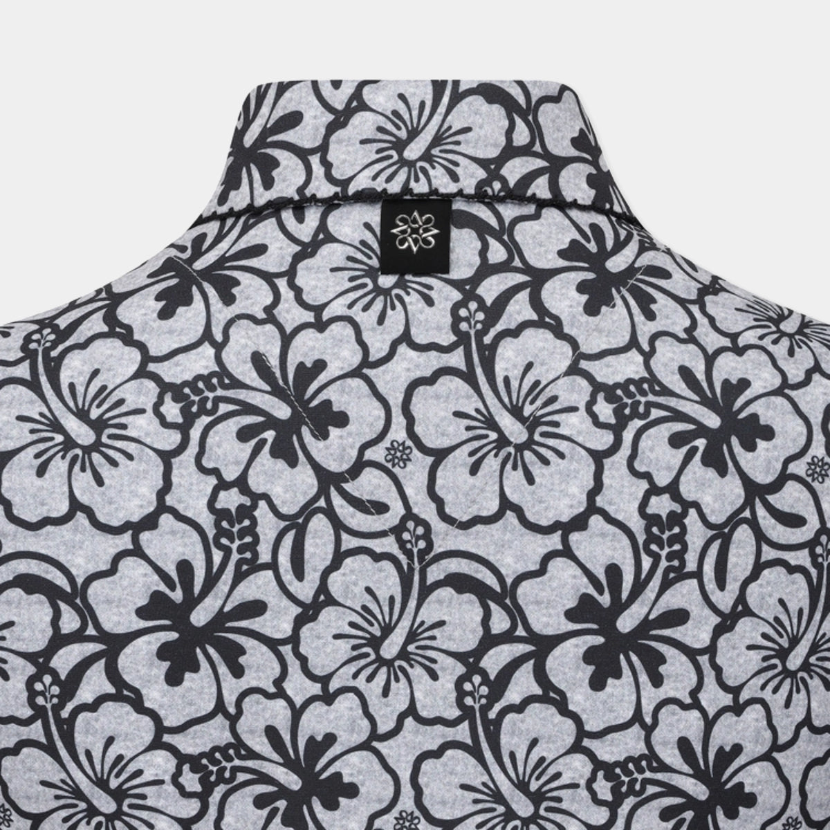 Close-up of the collar and upper back of a short-sleeve button-up shirt with a black hibiscus floral pattern on a light gray heathered fabric, featuring a small black brand tag with a white flower logo.
