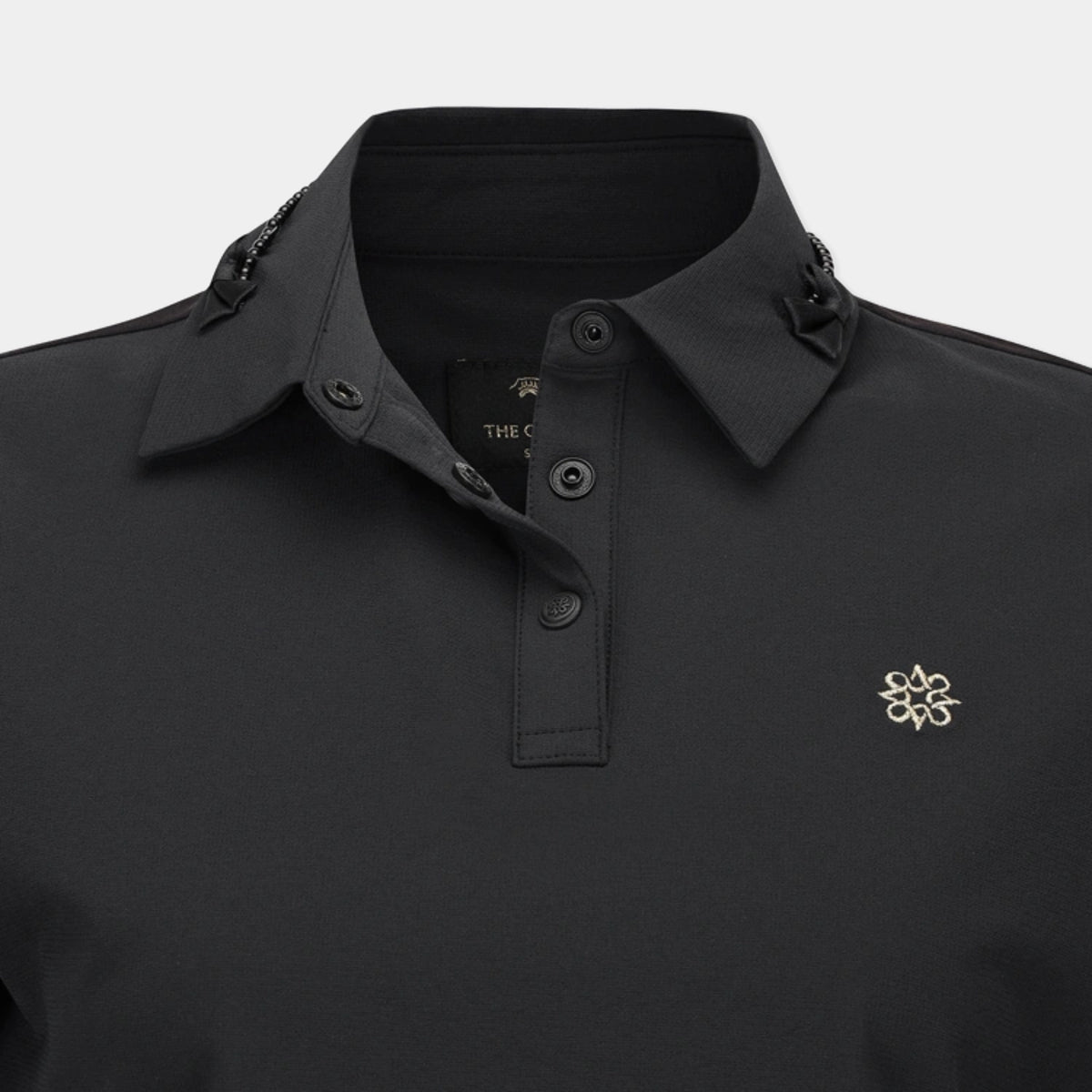 Close-up of a black polo shirt with a pointed collar, three-button placket, decorative chain detail on the collar, and a small gold embroidered logo on the left chest.