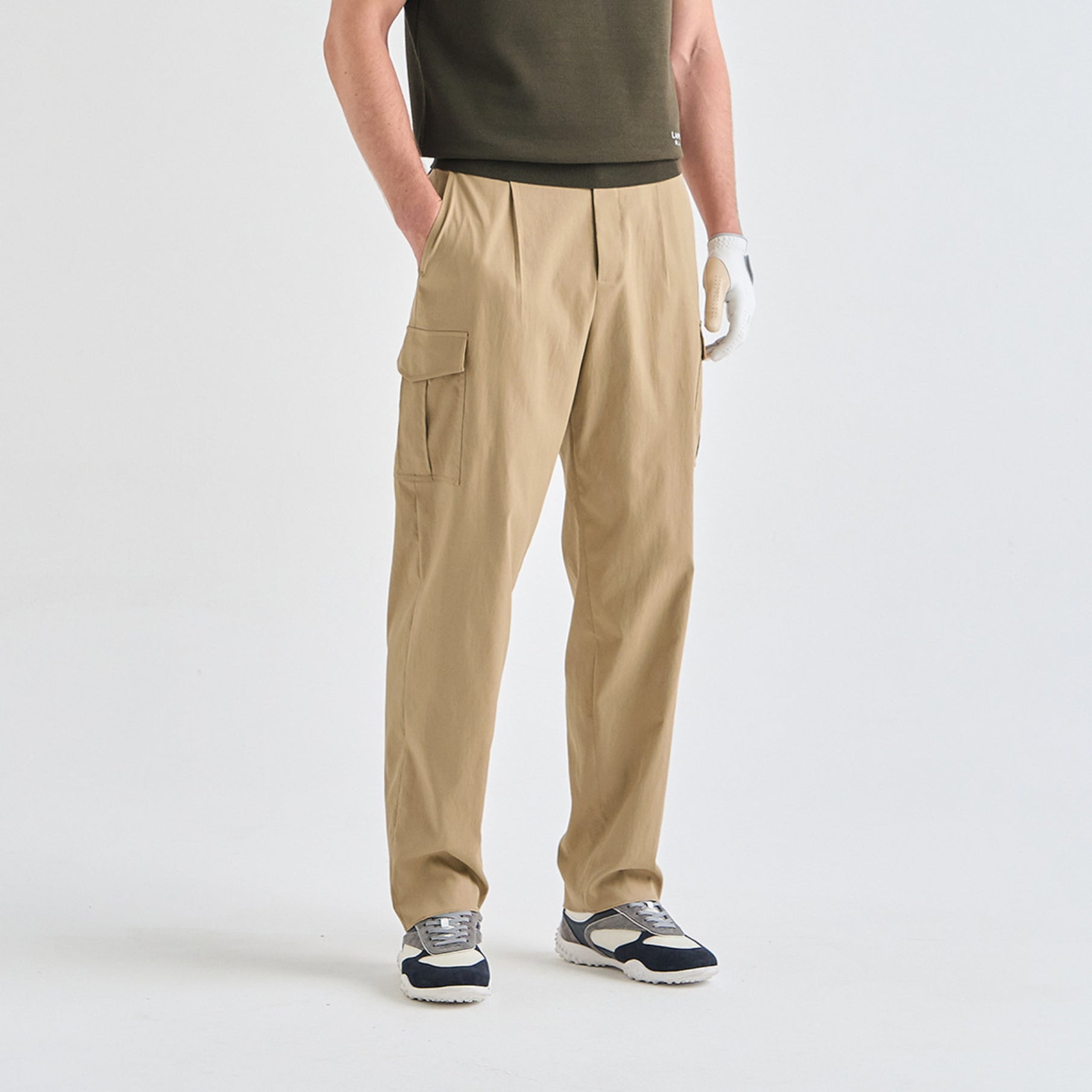 A person wearing khaki cargo pants, a dark olive green shirt, a white glove, and navy and white sneakers, standing against a light gray studio background.
