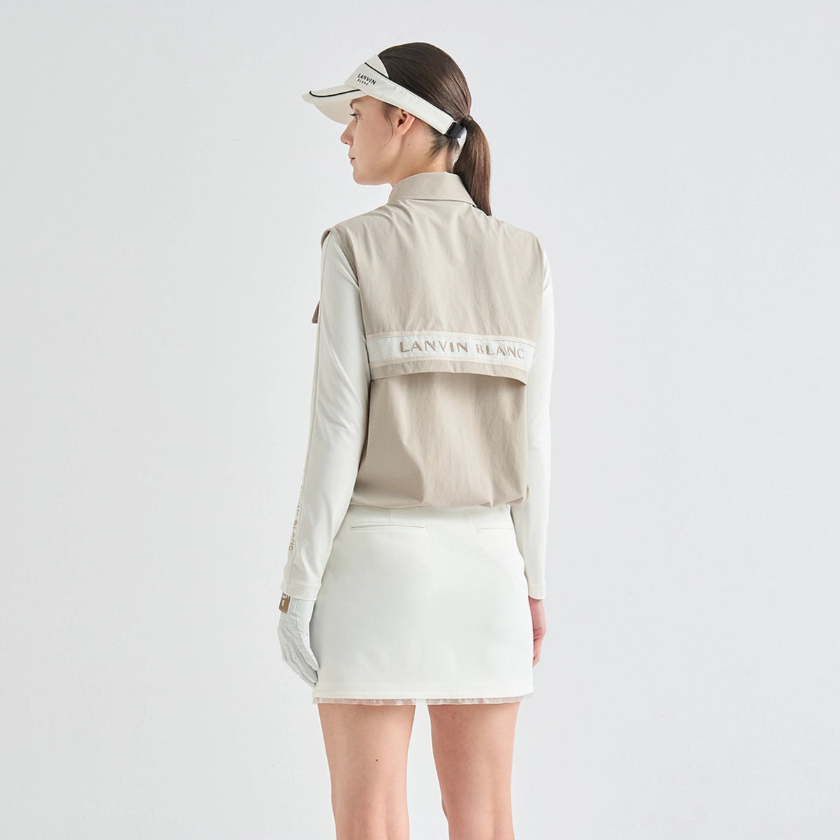 A woman shown from the back wearing a light beige sleeveless athletic vest over a white long-sleeve shirt, paired with a white mini skirt and visor. The vest features a horizontal band across the upper back with 'LANYAN ISLAND' printed on it.