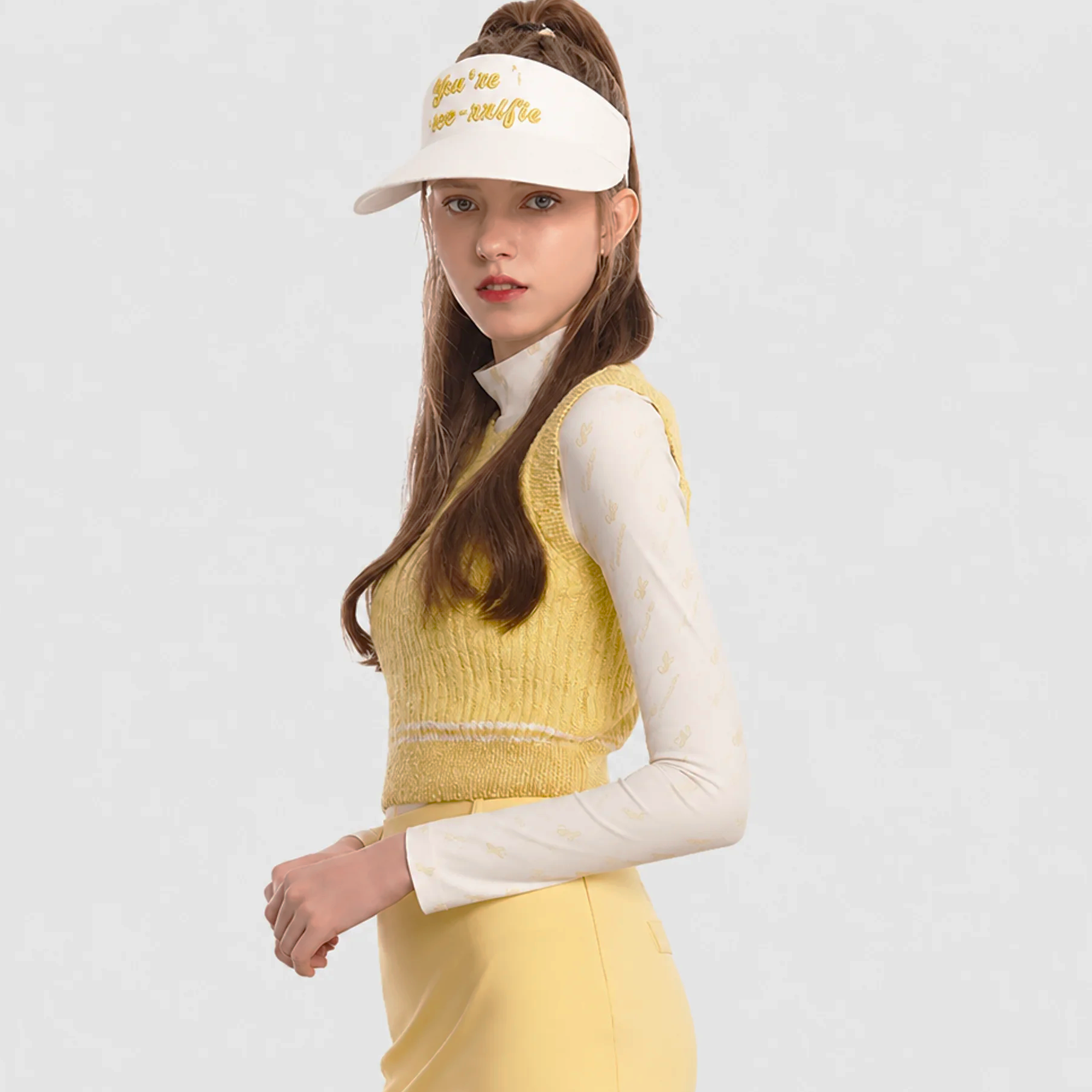 A woman wearing a white visor with gold embroidered text, a yellow knit sweater vest over a white long-sleeve top, and yellow pants, posed against a light gray studio background.