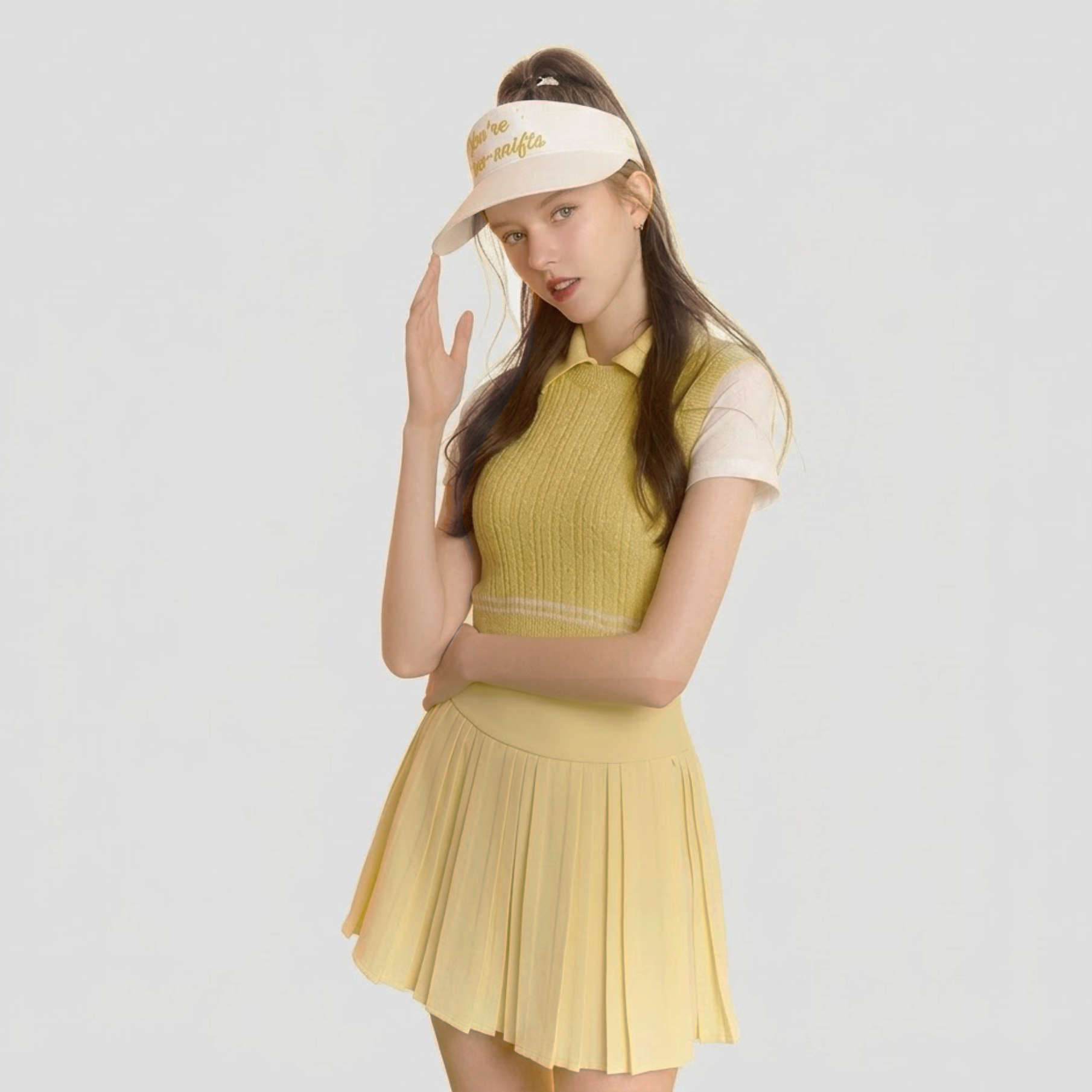 A woman modeling a yellow knit polo shirt and matching pleated tennis skirt set, paired with a cream visor, against a light gray studio background.