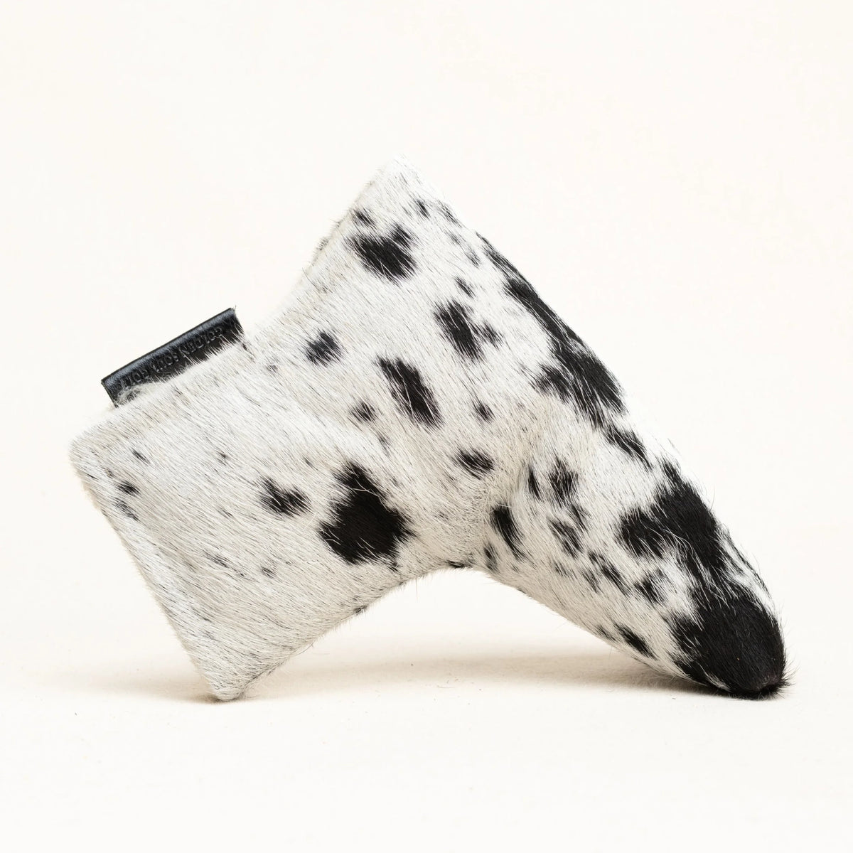 A golf club head cover made of black and white spotted cowhide, shown on a clean off-white background.