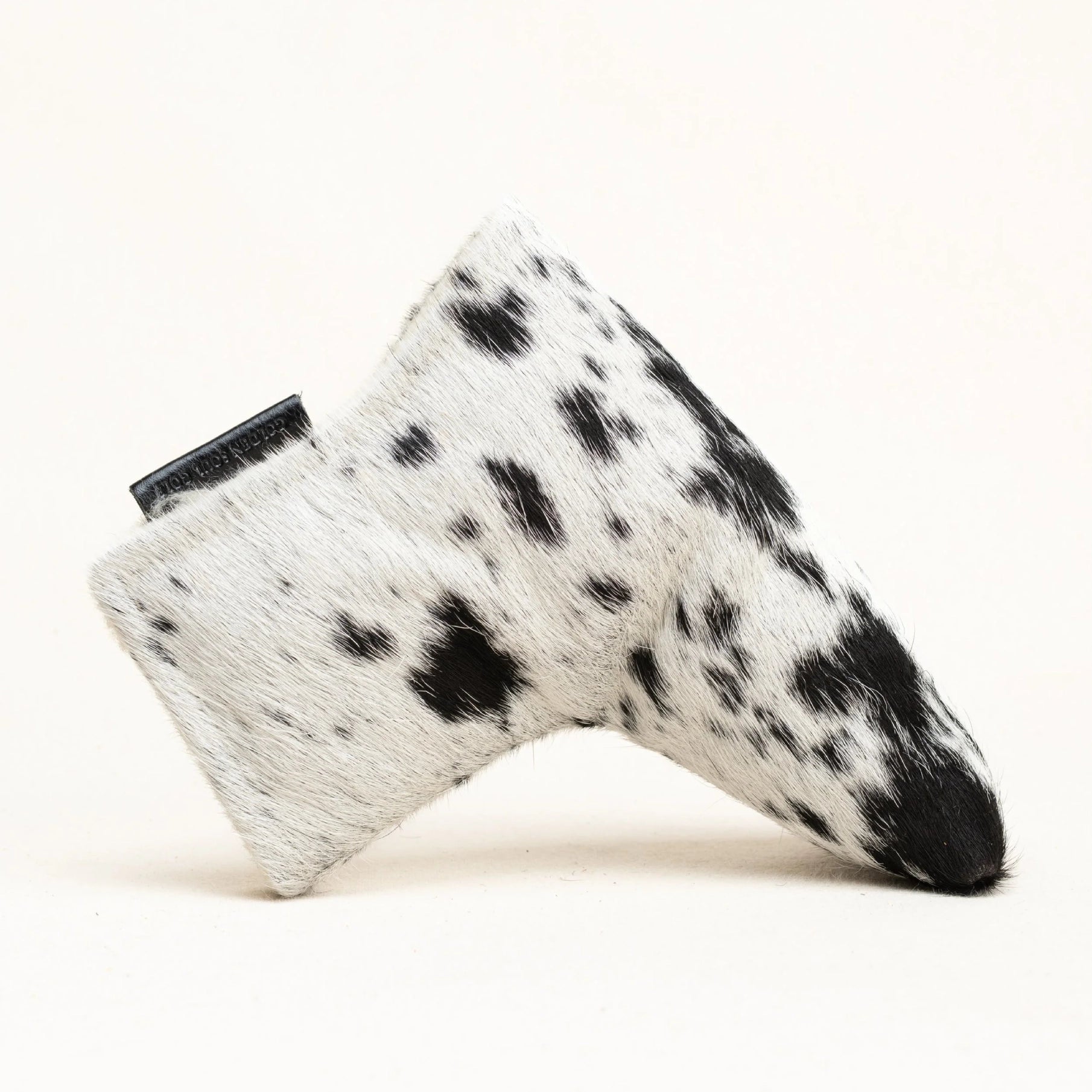 A golf club head cover made of black and white spotted cowhide, shown on a clean off-white background.