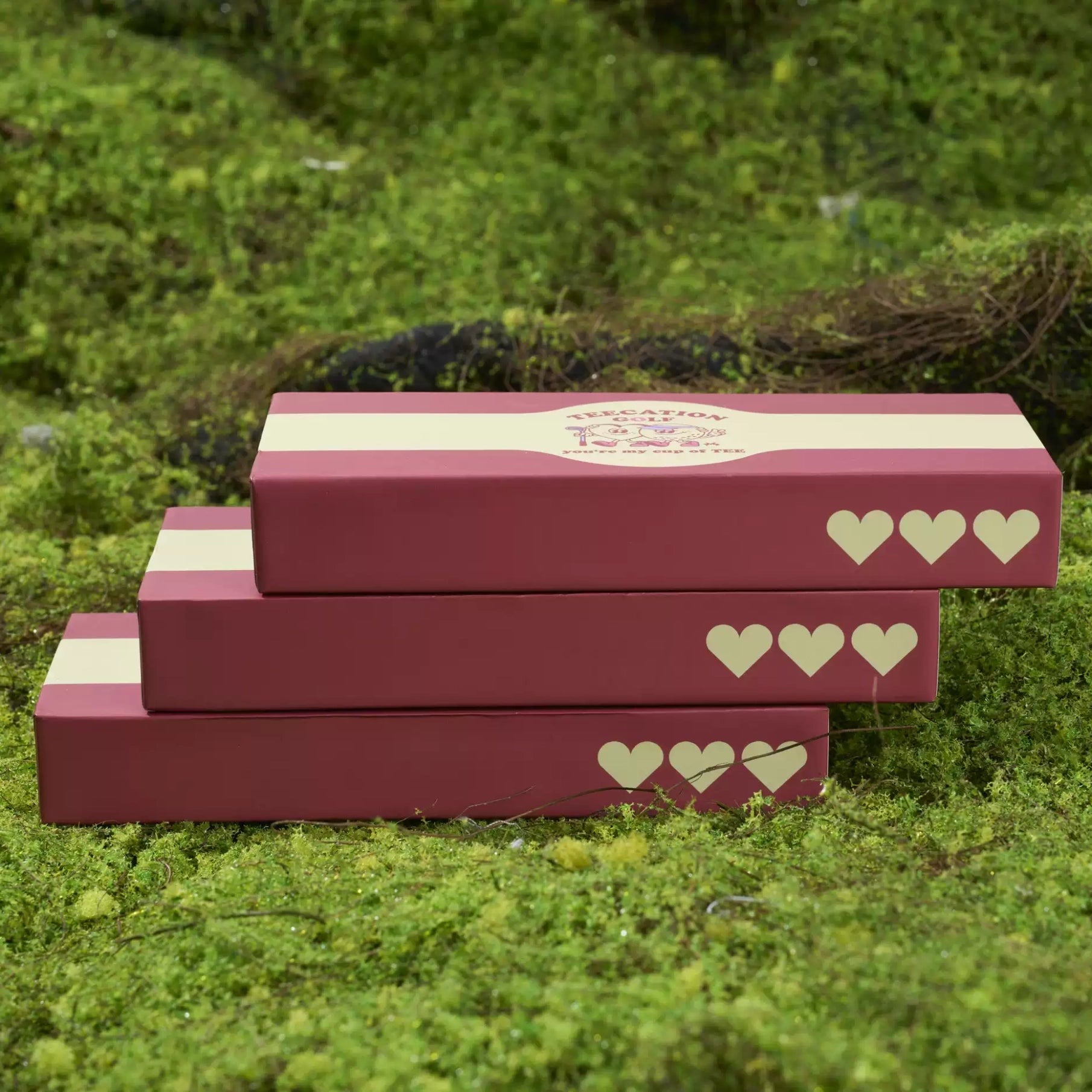 Three stacked rectangular gift boxes in dusty rose pink with cream horizontal bands and cream heart motifs, placed on a bed of green moss.