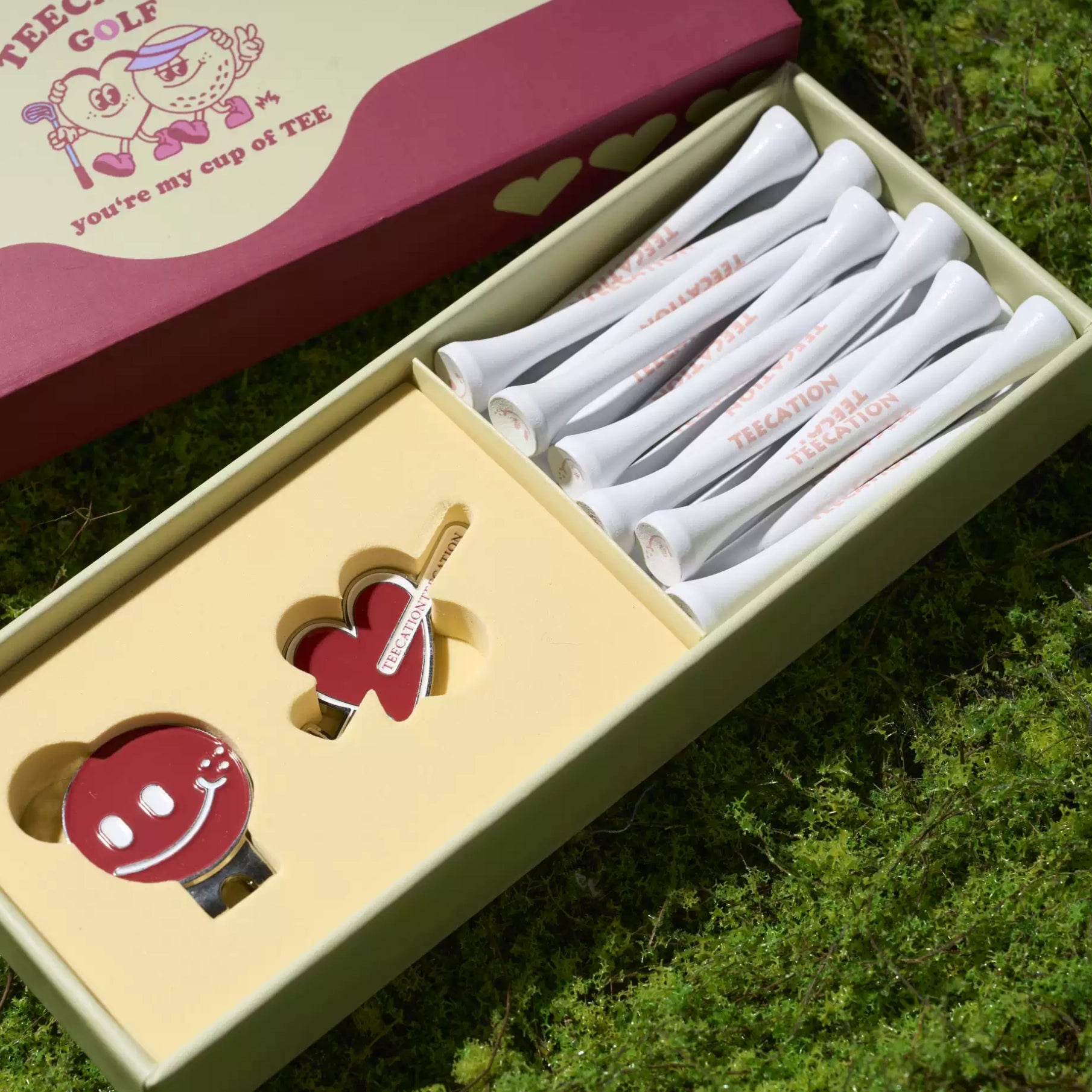 A boxed golf gift set placed outdoors on green grass, showing a red and cream presentation box with a set of white golf tees and a red teedie iron.